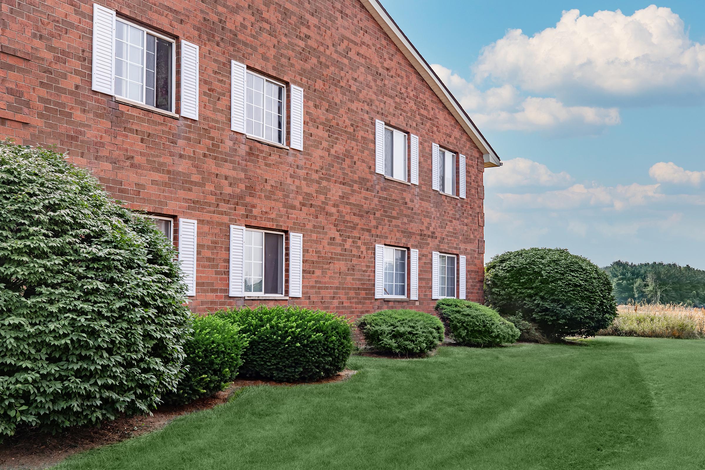 A brick building with white-trimmed windows, surrounded by neatly manicured bushes and a grassy lawn, under a blue sky with fluffy clouds.