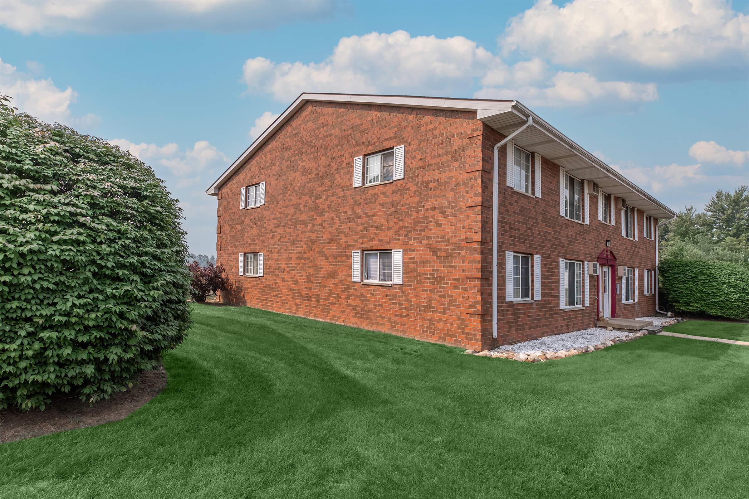 A brick apartment building with multiple windows, surrounded by a well-maintained green lawn and shrubs. The building features a red entrance door and is set against a backdrop of a partly cloudy sky.