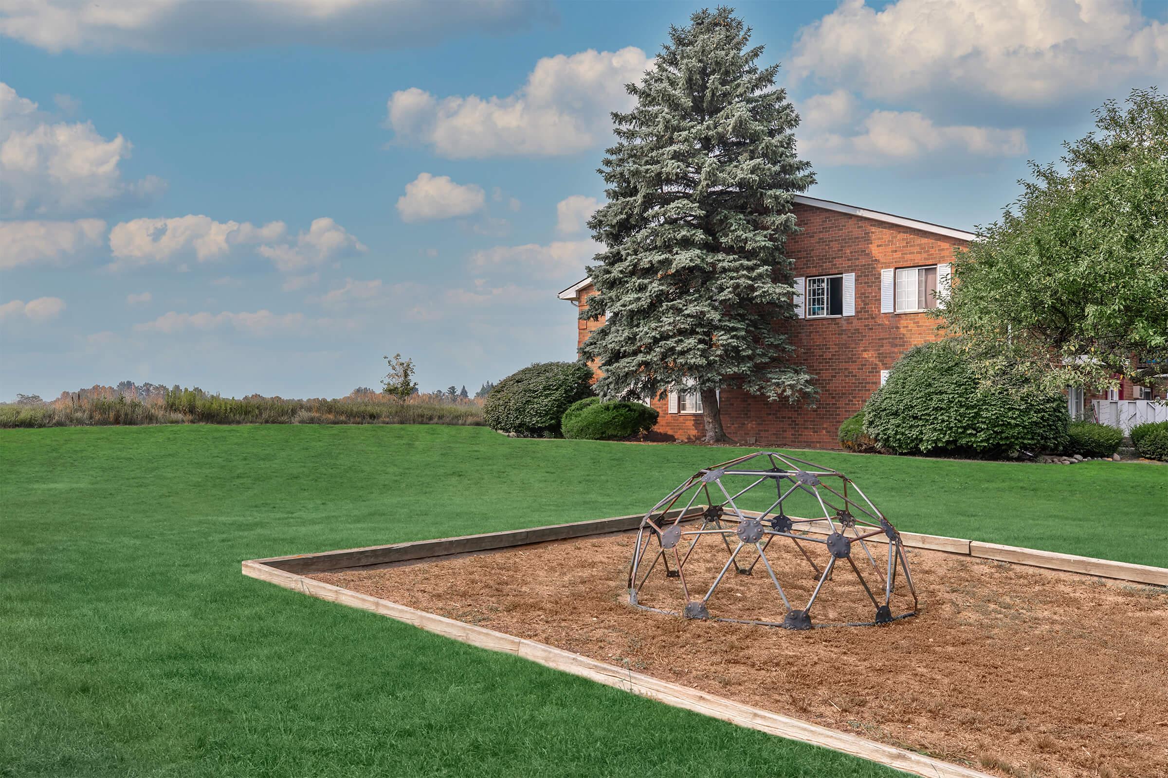 A play area featuring a geometric climbing structure on dry, sandy ground, surrounded by green grass. In the background, a brick building with multiple windows and a large evergreen tree can be seen, under a cloudy sky.