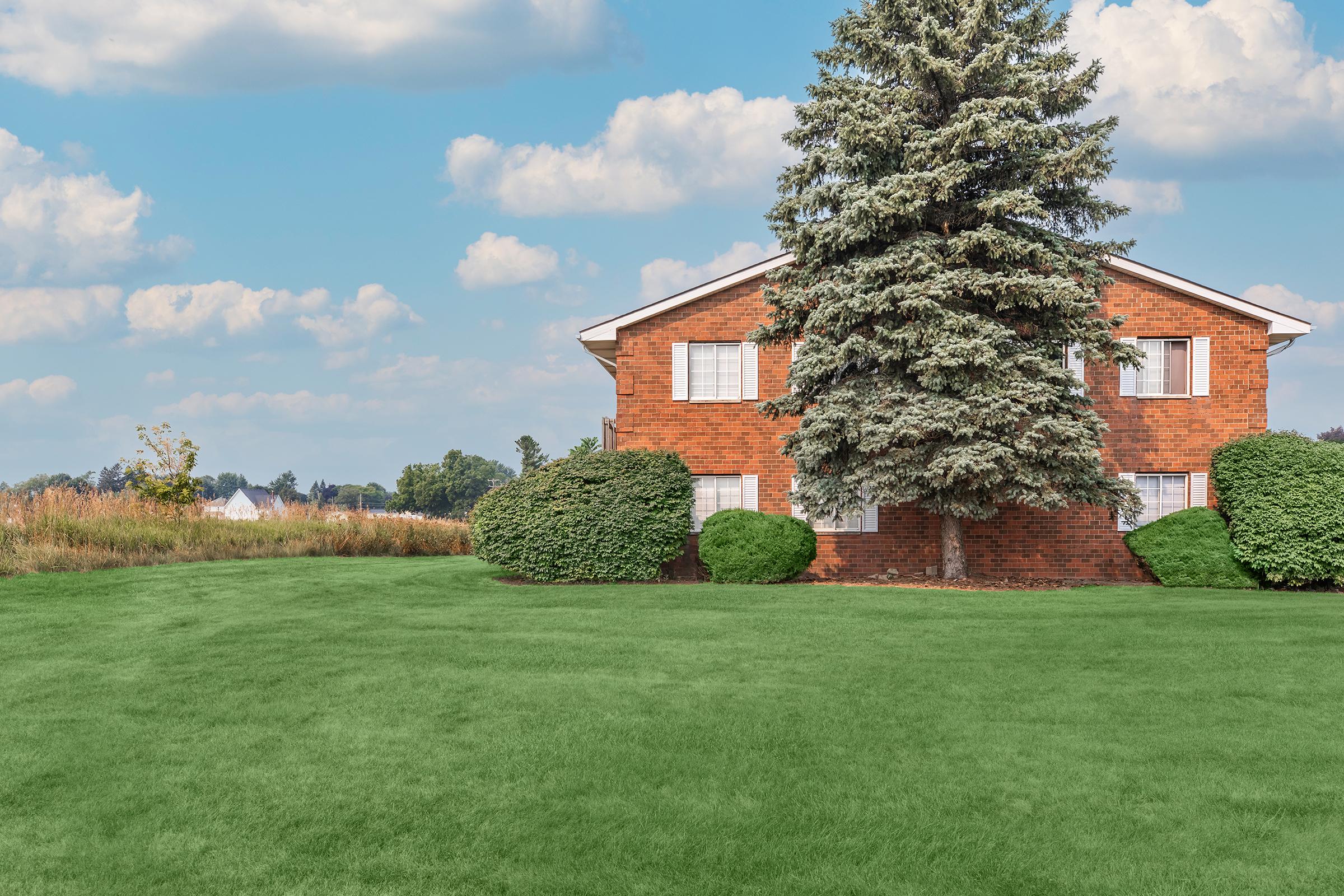 A well-maintained, two-story brick house with large windows, surrounded by neatly trimmed bushes and a tall evergreen tree in the yard. A green lawn stretches in front of the house, with a clear blue sky and fluffy clouds overhead. Fields are visible in the background.