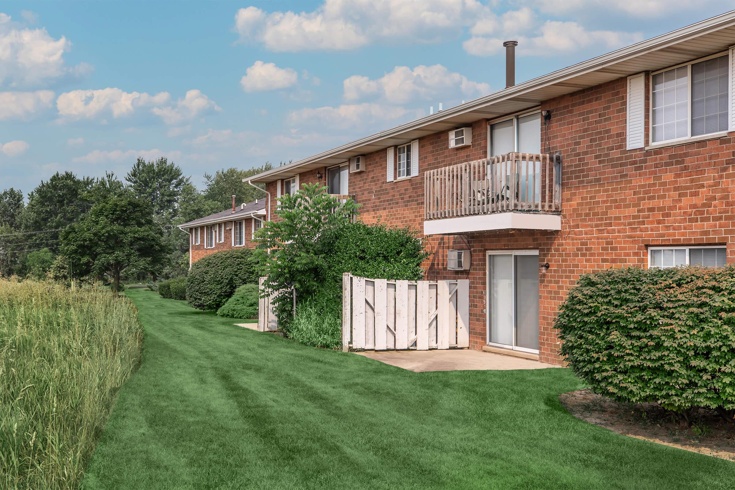 A row of brick apartment buildings with white balconies, surrounded by well-maintained green grass and shrubs. A pathway runs alongside the buildings, leading to the entrance of each unit. The sky is partially cloudy, indicating a pleasant day.