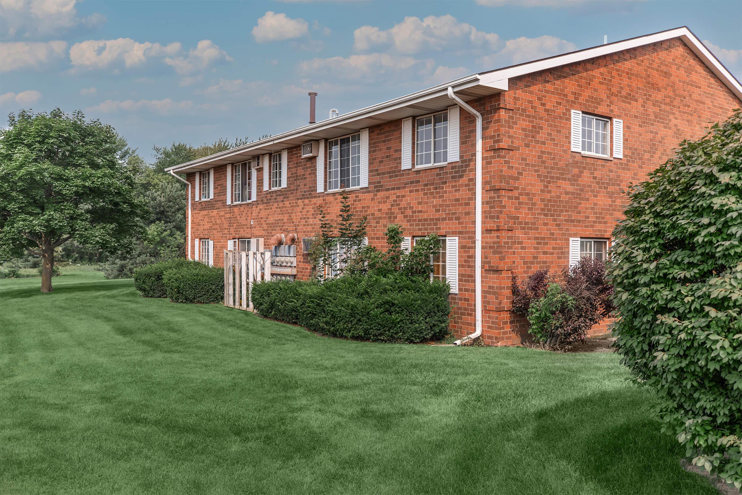 A brick residential building with multiple windows and a well-maintained lawn. There are trees and shrubs surrounding the property, creating a lush green landscape. The sky is partly cloudy, adding a serene atmosphere to the scene.