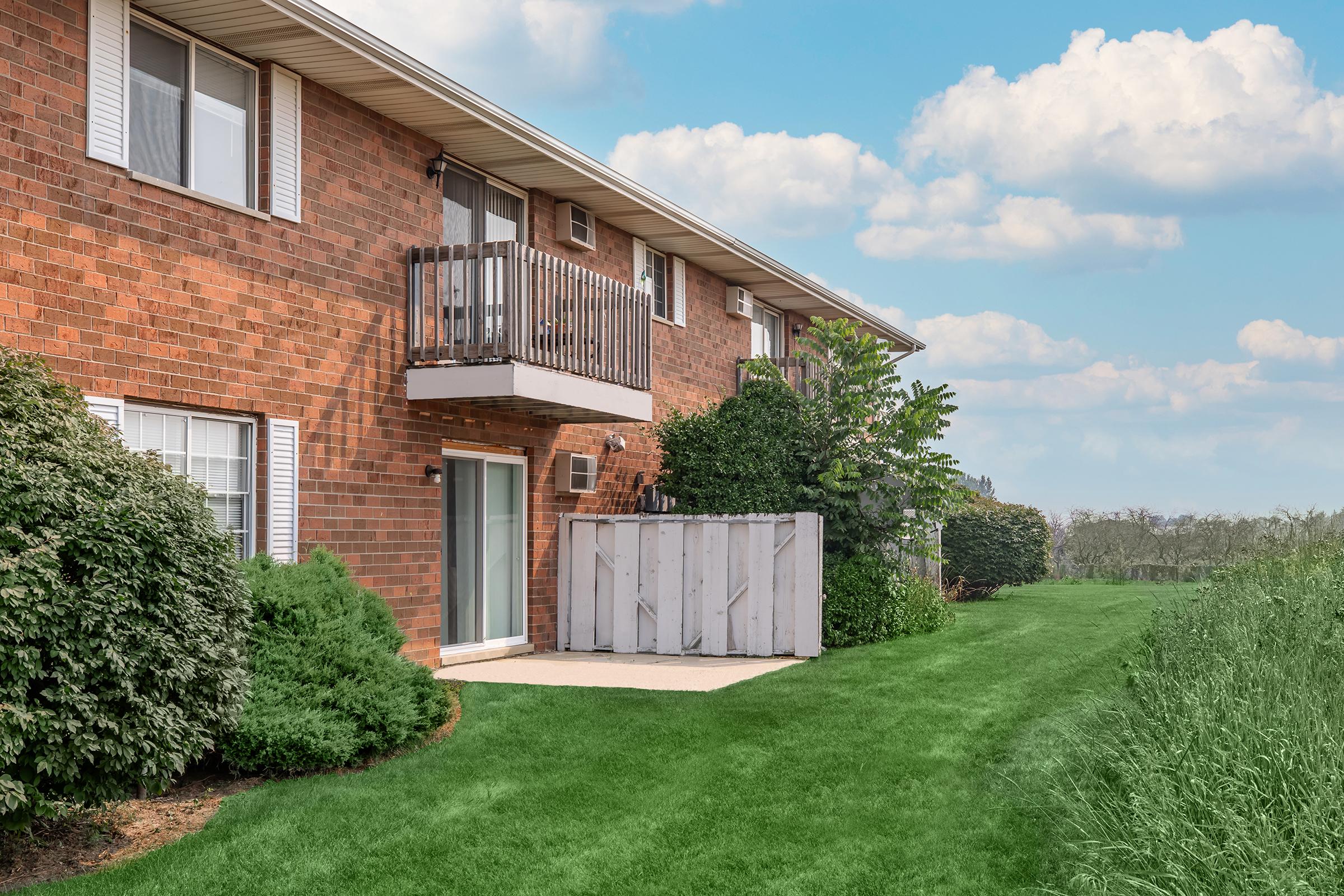 A view of an apartment building with brick siding, featuring a balcony, air conditioning unit, and a fenced patio area. The surrounding landscape includes lush green grass and neatly trimmed bushes, with a clear blue sky and fluffy clouds in the background.