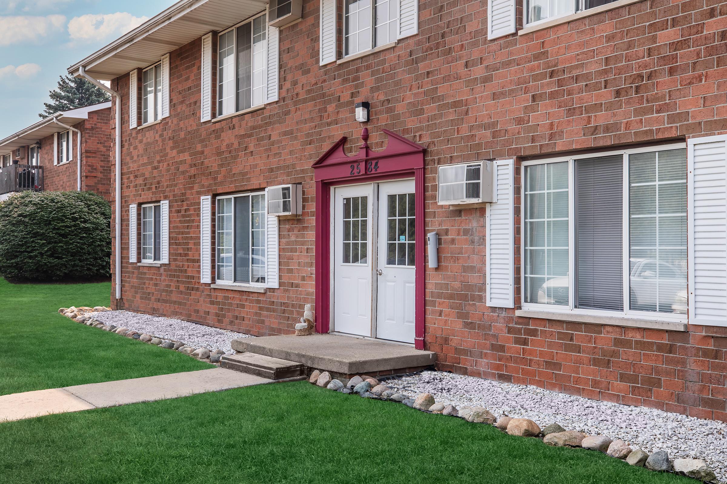 A brick apartment building entrance featuring a white double door framed by decorative trim. Windows on either side allow natural light, and small air conditioning units are mounted on the lower windows. A neatly maintained lawn with gravel landscaping and decorative stones leads to the entrance.