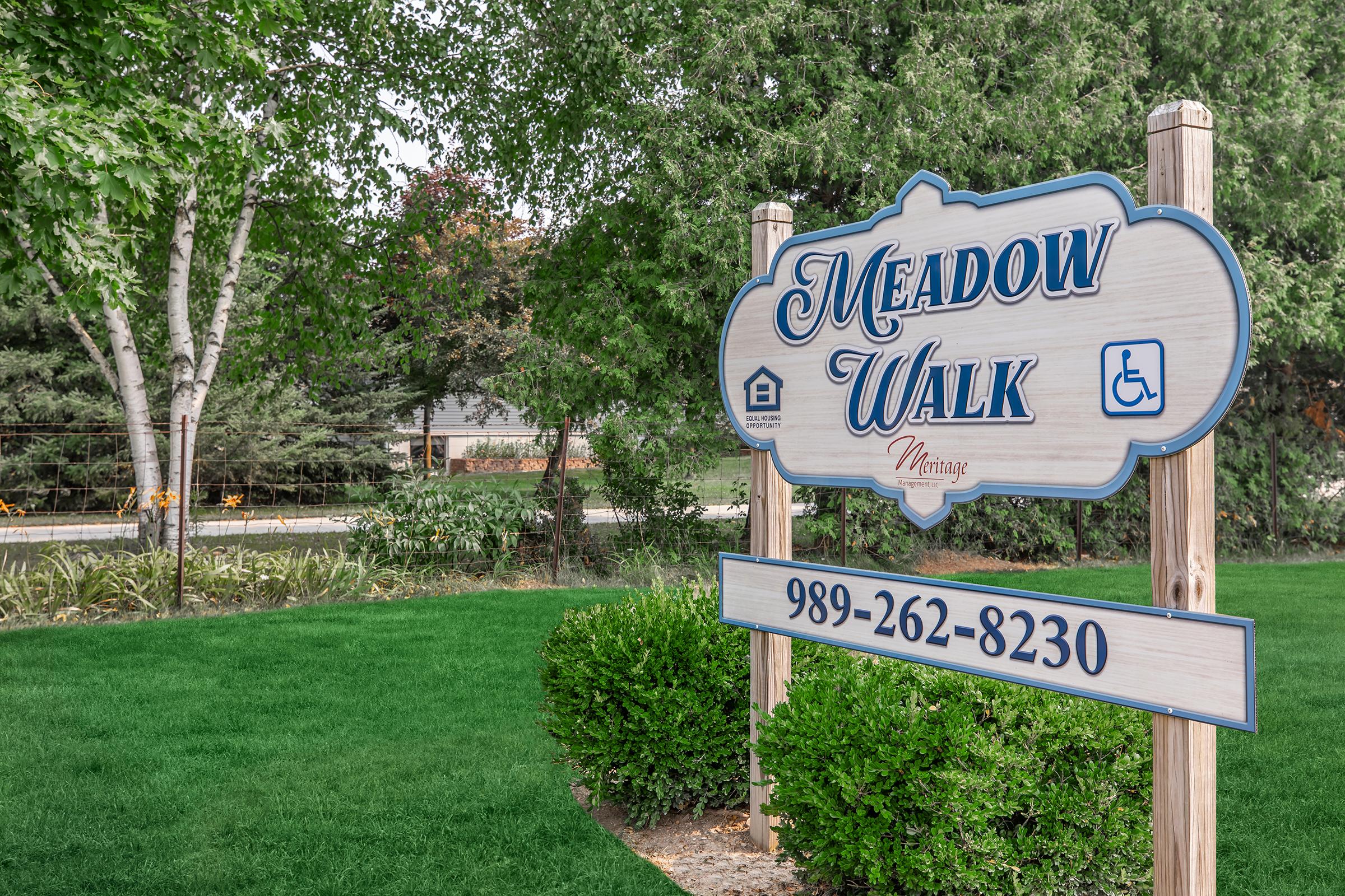 Sign for "Meadow Walk" featuring a decorative blue and white design. The sign includes a house icon and accessibility symbol, along with a phone number (989-262-8230). The surrounding area has green grass and trees, creating a welcoming atmosphere.