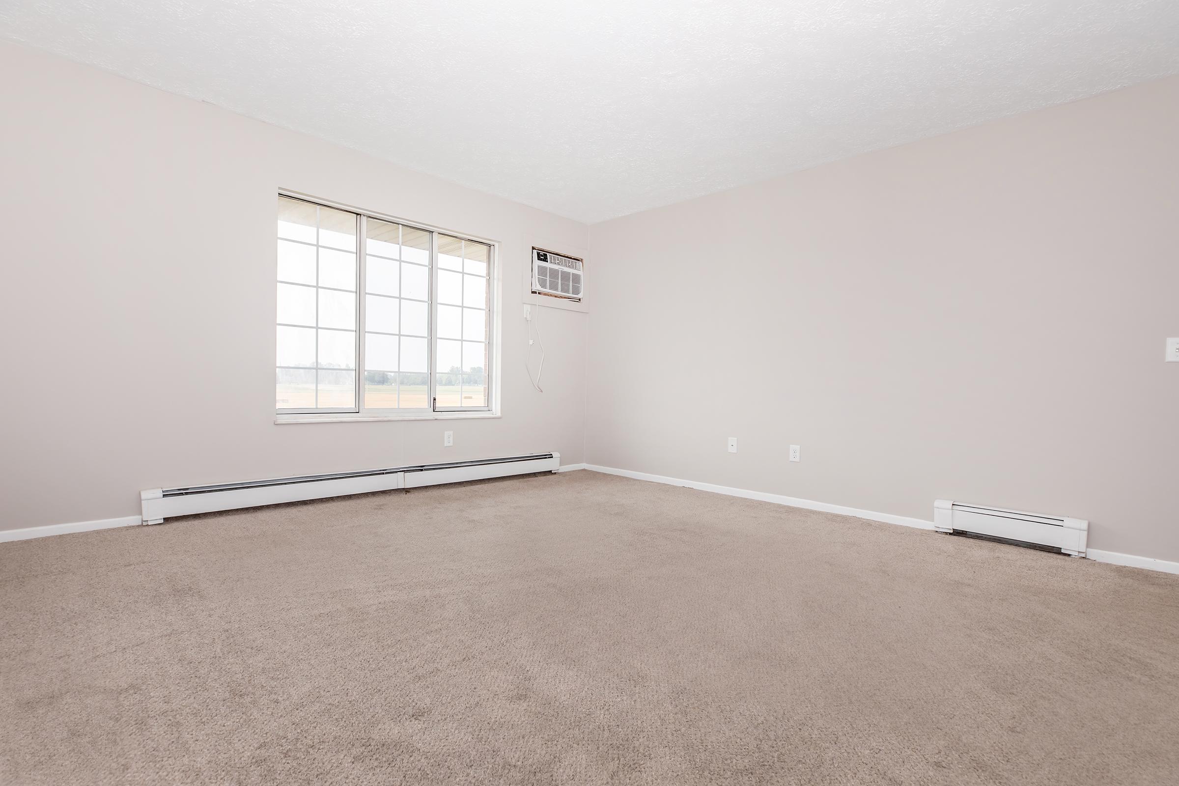 A spacious, empty room featuring beige carpet and light gray walls. A large window allows natural light to fill the space, with an air conditioning unit mounted above. Baseboard heating can be seen along two walls, creating a simple and clean aesthetic suitable for various uses.