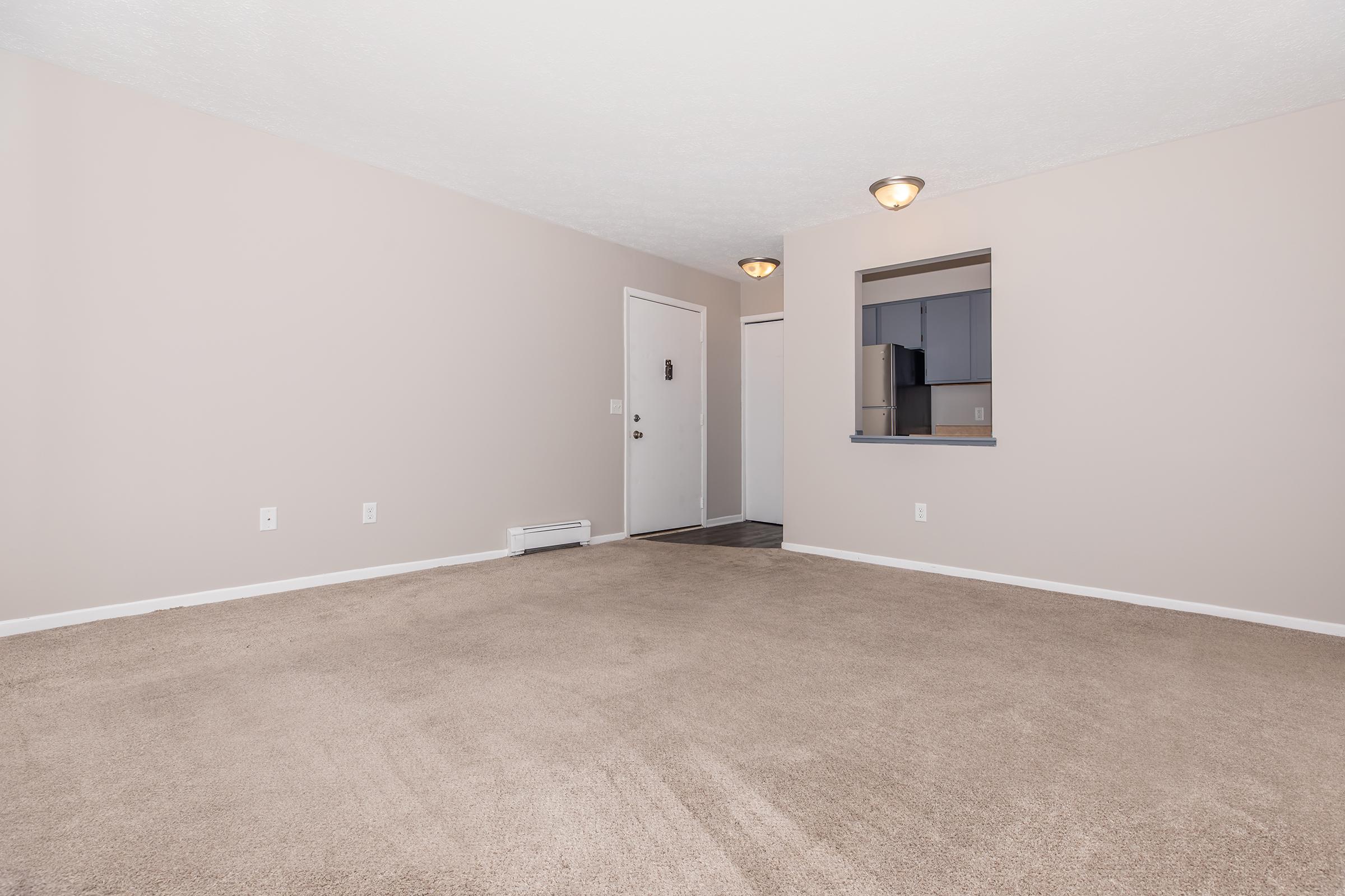 Empty living room with beige carpet and light beige walls. A front door is visible on the left, and there is an opening to a kitchen on the right with a dark countertop. Two light fixtures hang from the ceiling, providing soft lighting. The space appears clean and spacious, ready for furnishing.