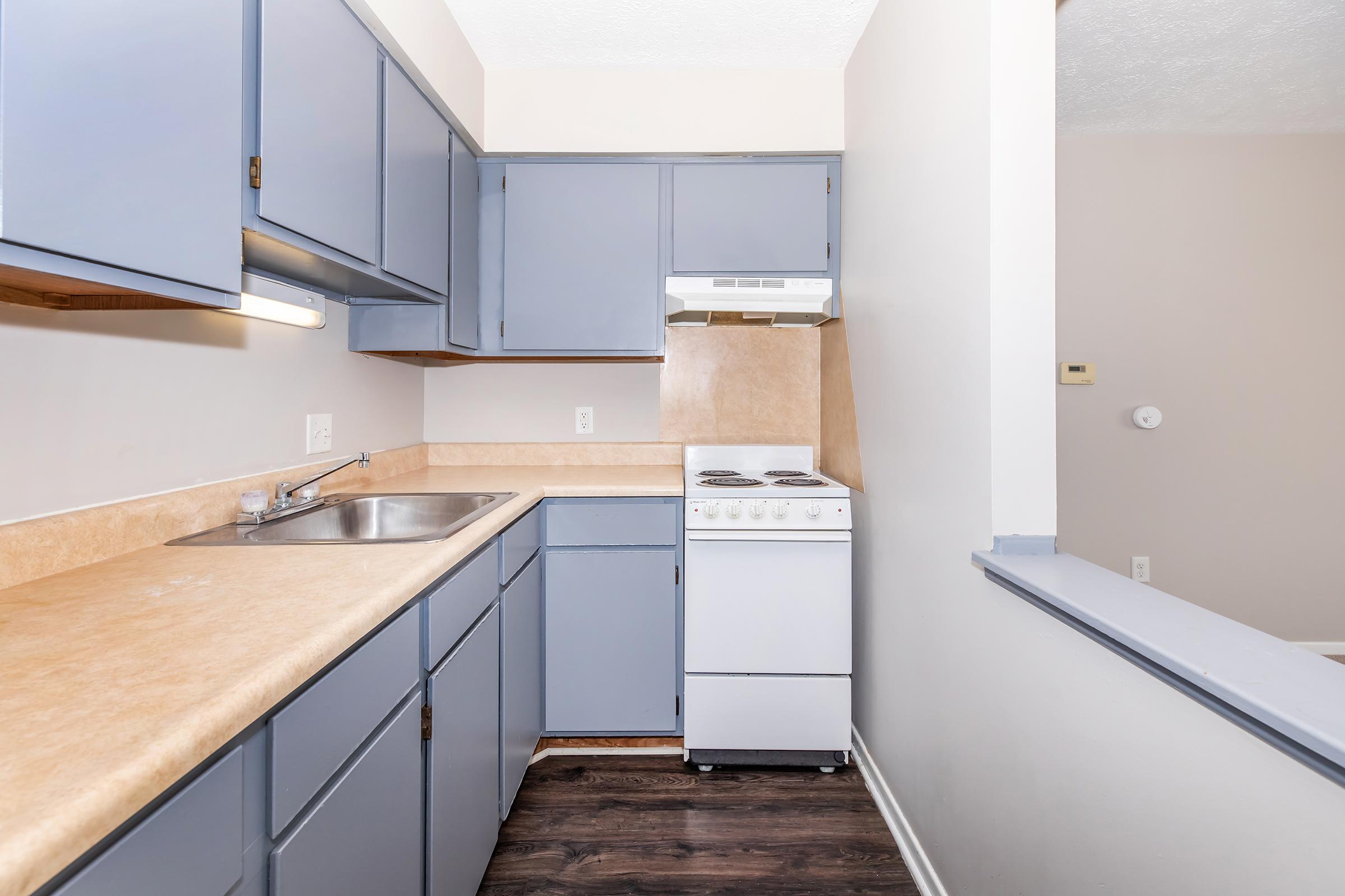 A small kitchen space featuring light blue cabinets, a beige countertop, a stainless steel sink, and a white stove. The walls are painted in neutral colors, and there is a window above the sink for natural light. The flooring is dark, adding contrast to the overall design.