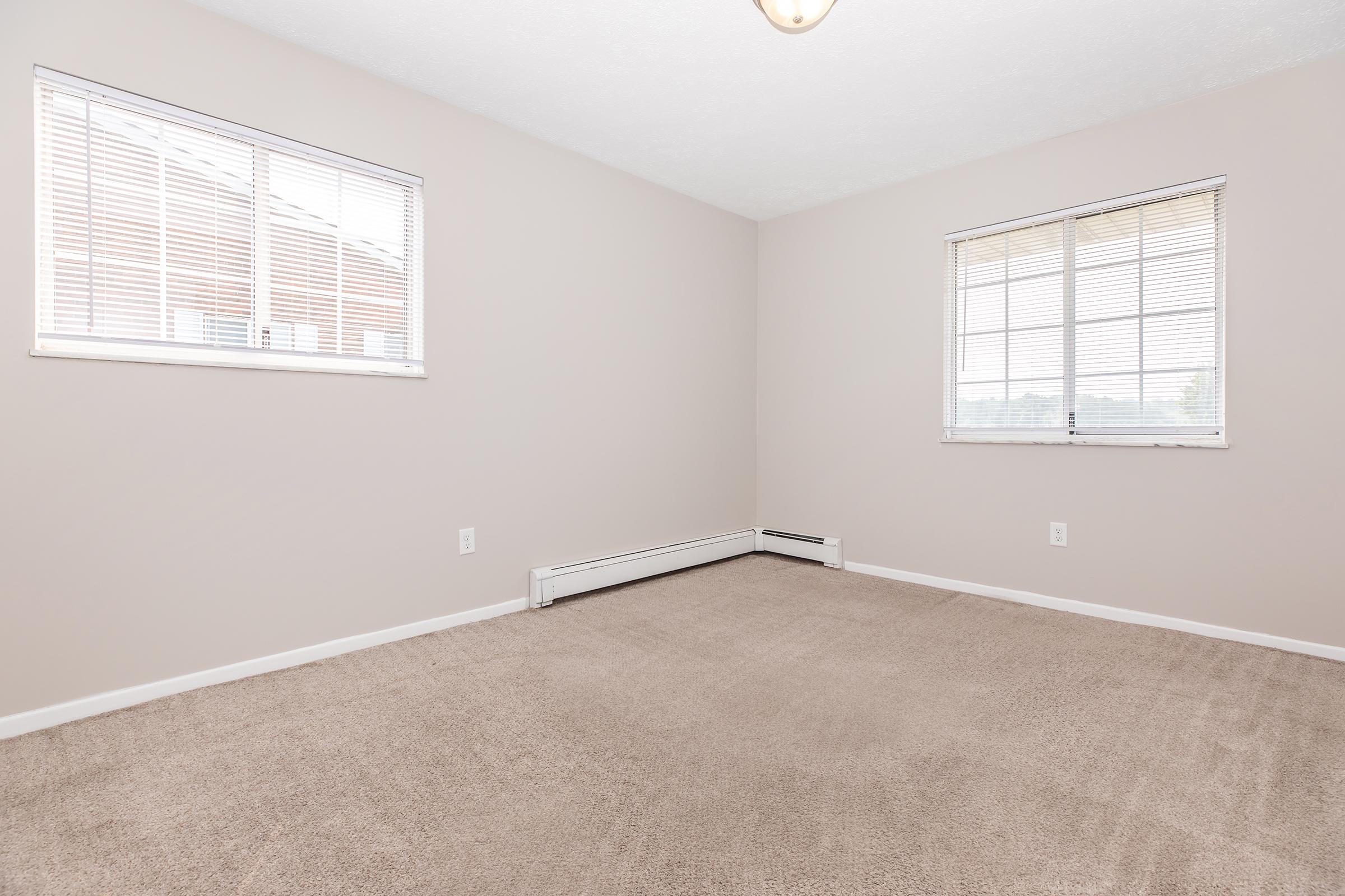 Empty room with light beige walls and carpet. Two windows allow natural light, with white blinds partially closed. A baseboard heater runs along the wall. The space is uncluttered and unfurnished, emphasizing the clean and airy feel of the room.