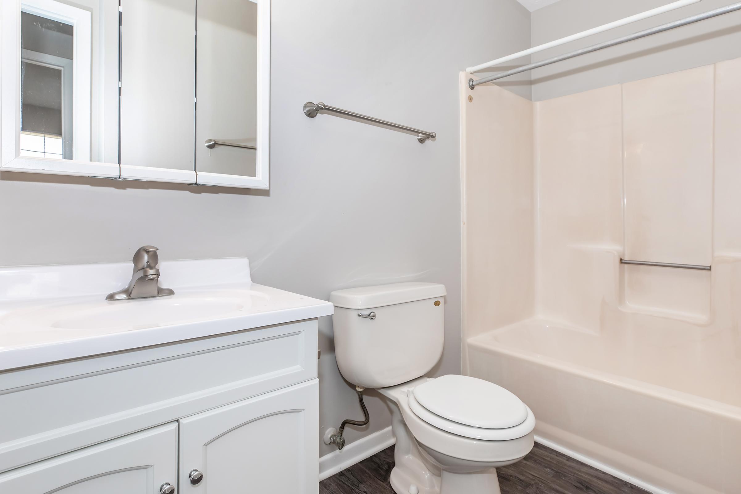 A well-lit bathroom featuring a white sink with a mirror above, a toilet, and a beige bathtub with a shower rod. The walls are painted light gray, and the flooring is dark. The space is clean and modern, highlighting simple fixtures and neutral colors.