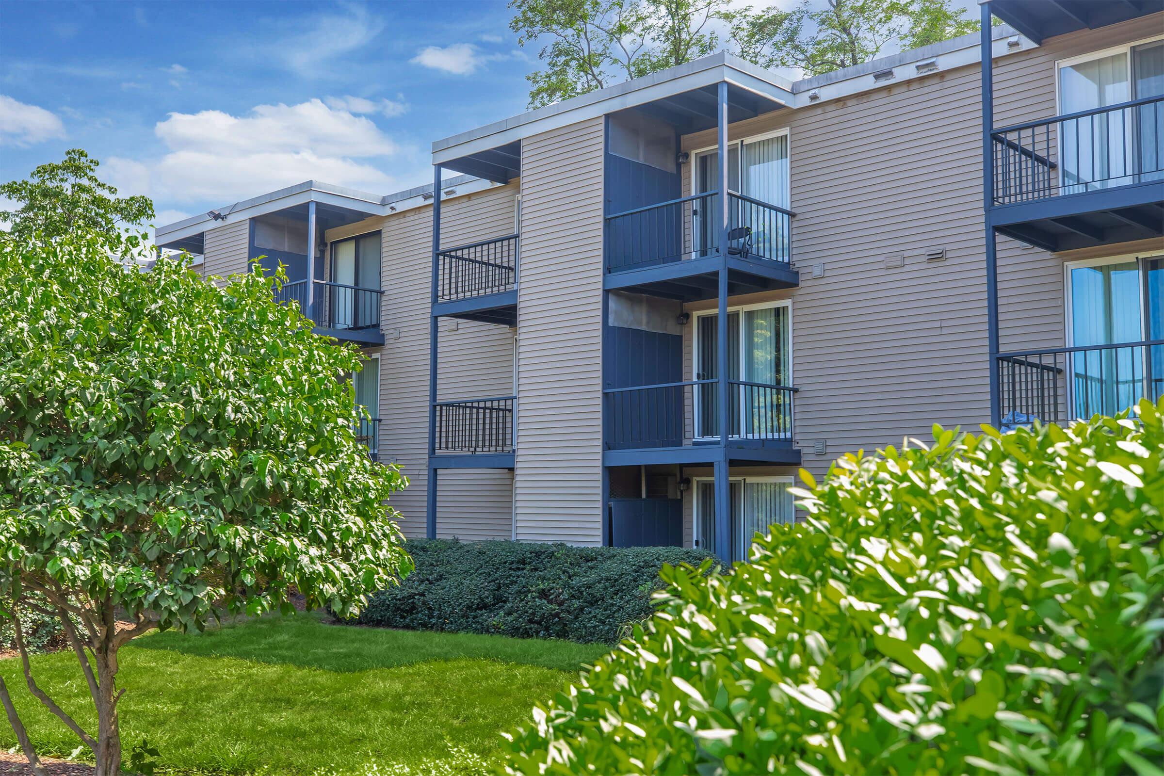 A modern multi-story apartment building with balconies, surrounded by green shrubbery and grass. The building features light-colored siding and large windows. A blue sky with a few clouds is visible in the background, creating a bright and inviting atmosphere.