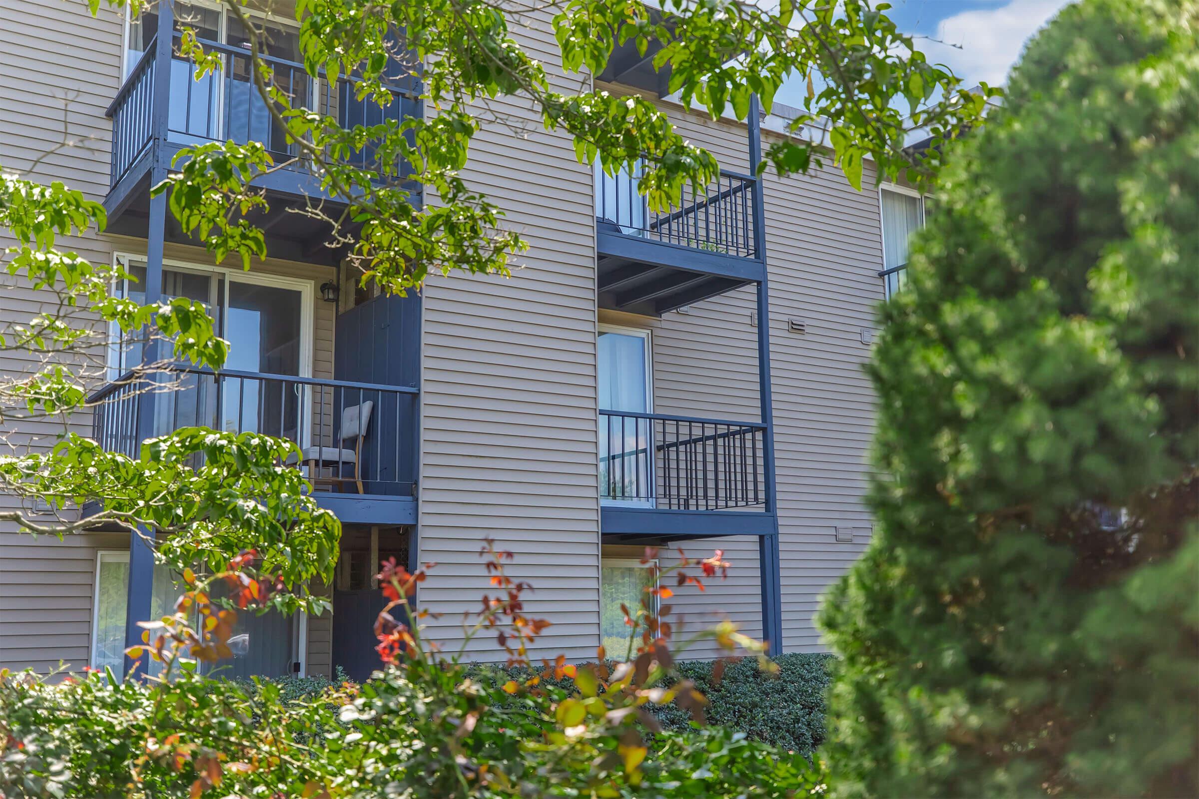 A view of a multi-story residential building with balconies, framed by green foliage and shrubs in the foreground. The building has light-colored siding and several windows, with blue railings on the balconies visible. The sky is clear, indicating a sunny day.