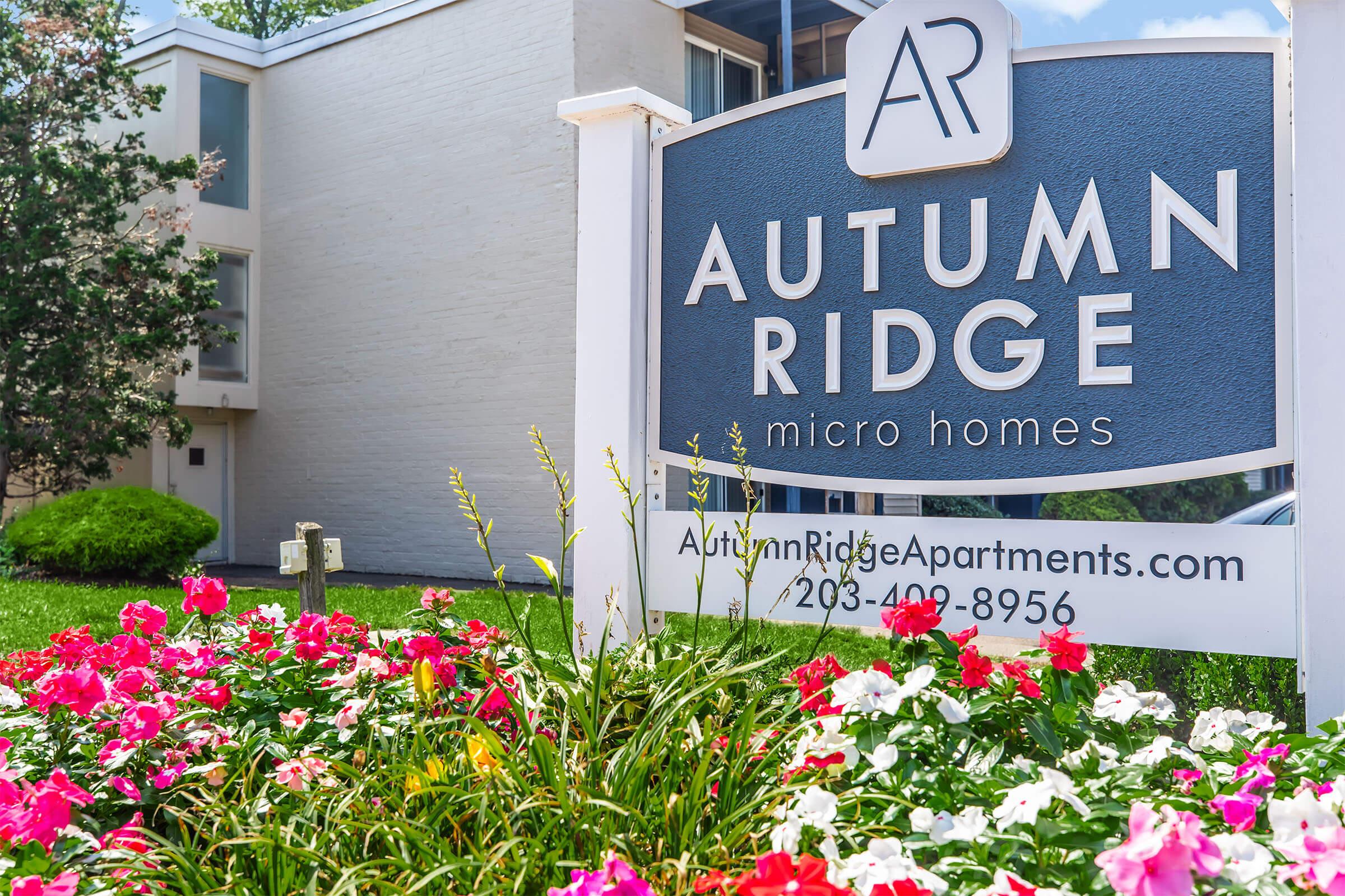 Sign for Autumn Ridge micro homes, featuring the logo "AR" at the top. The sign is surrounded by colorful flower beds with vibrant blooms. In the background, a building is partially visible, suggesting a residential area. The website and phone number for Autumn Ridge Apartments are also displayed.