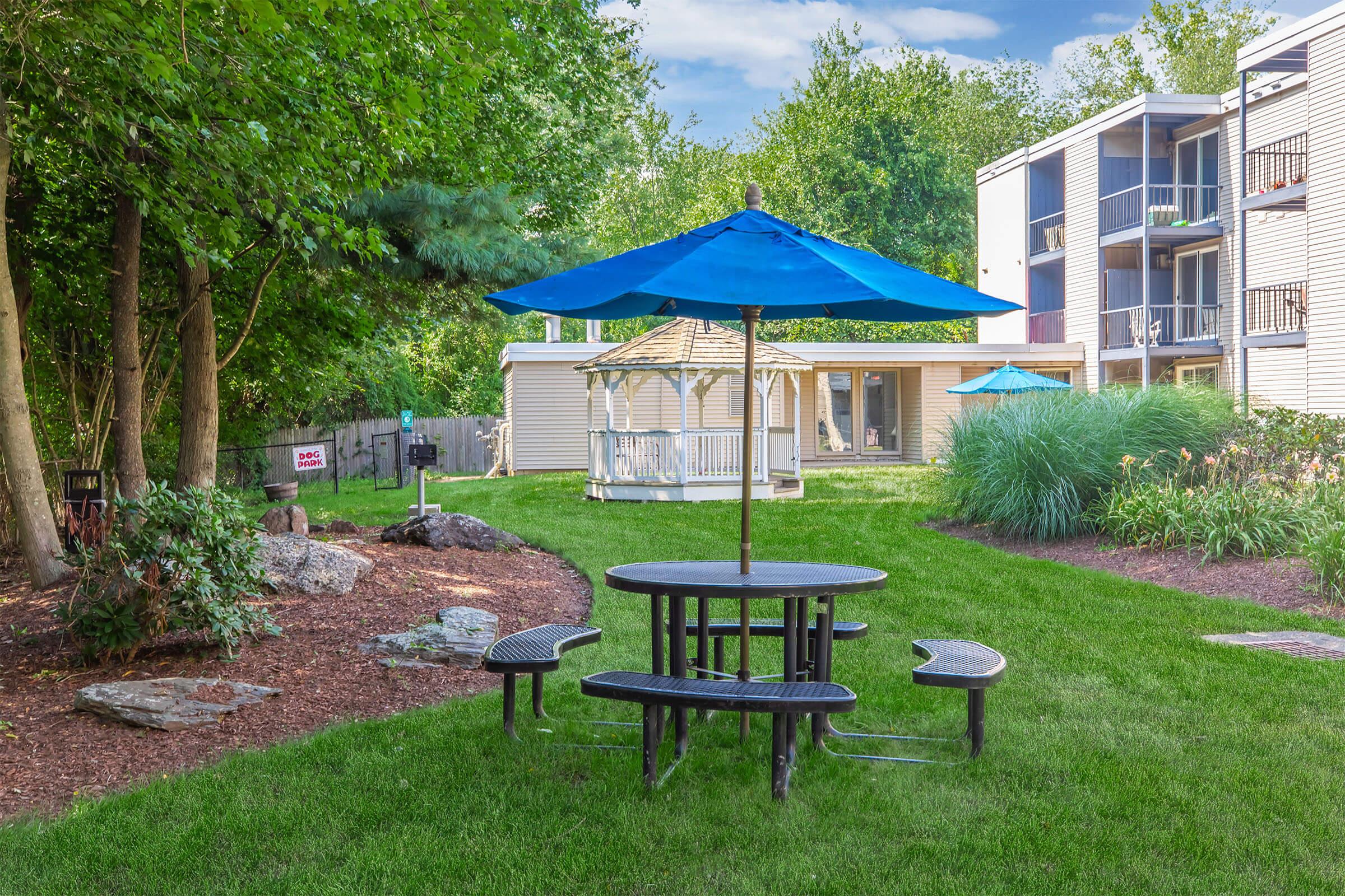 A grassy area features a black picnic table with attached benches, shaded by a bright blue umbrella. In the background, there is a white gazebo and a multi-story building with balconies. Lush green trees and landscaping surround the scene, creating a relaxing outdoor space.