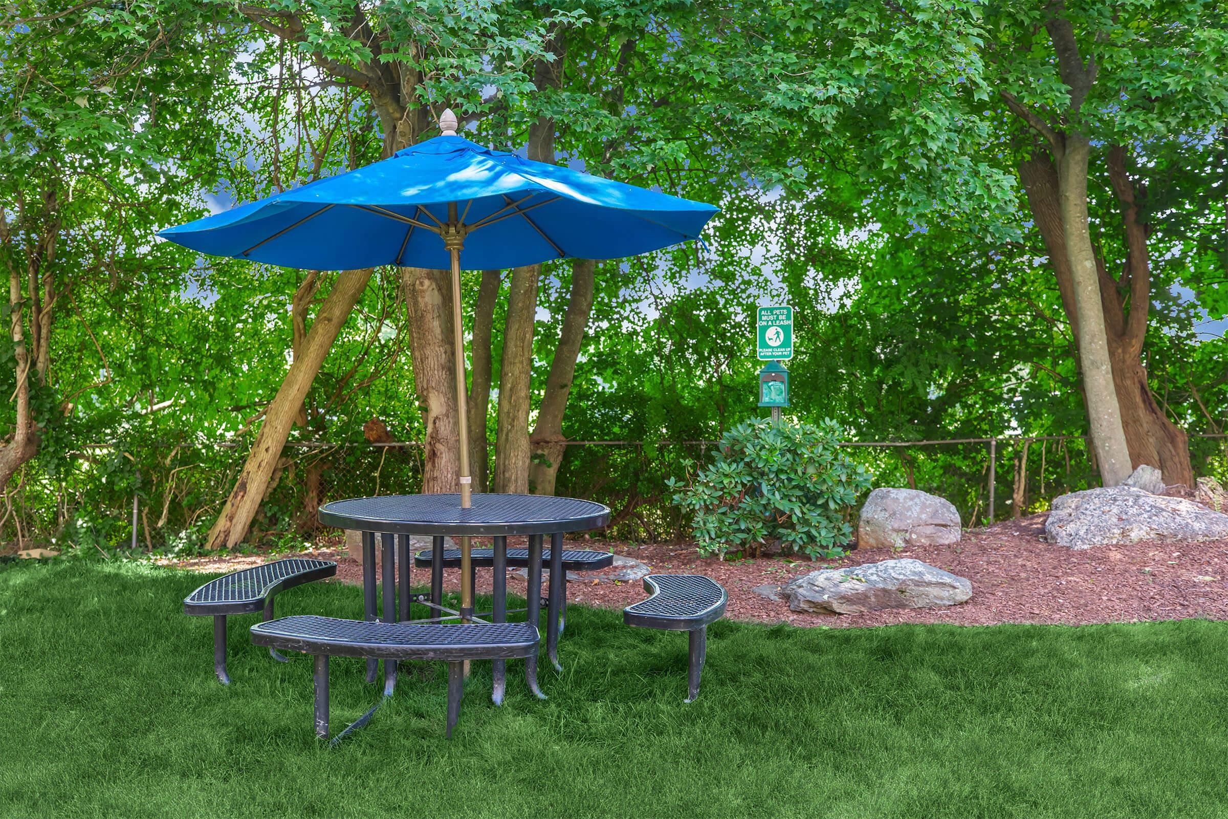 A round picnic table with attached benches sits under a large blue umbrella, surrounded by green grass and trees. A signpost is visible in the background, adding to the outdoor setting's inviting and serene atmosphere.