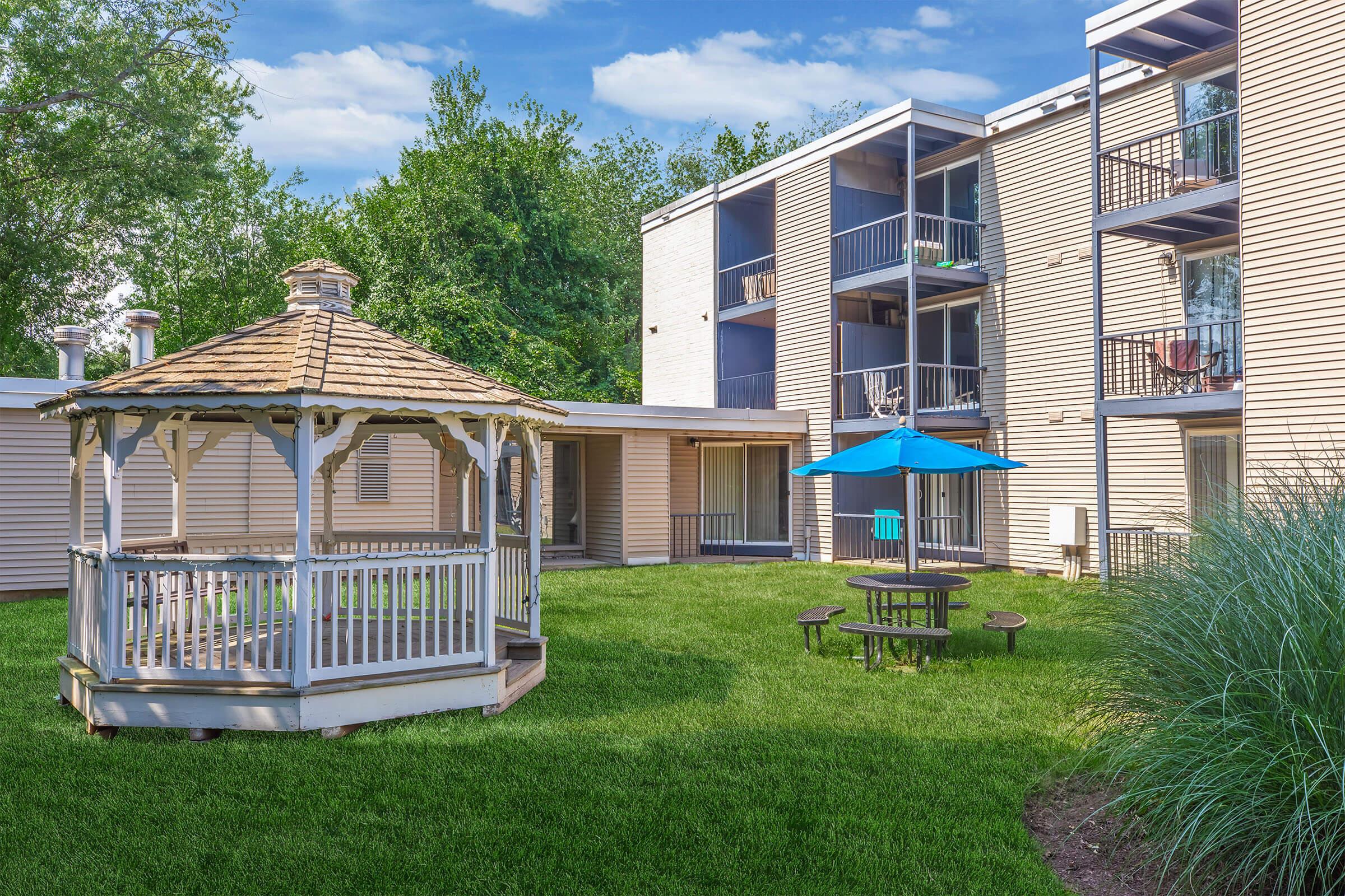 A well-maintained outdoor area featuring a gazebo with a shingled roof, a picnic table underneath a blue umbrella, and a grassy lawn. Surrounding the lawn are multi-story residential buildings with balconies and trees in the background under a clear blue sky.