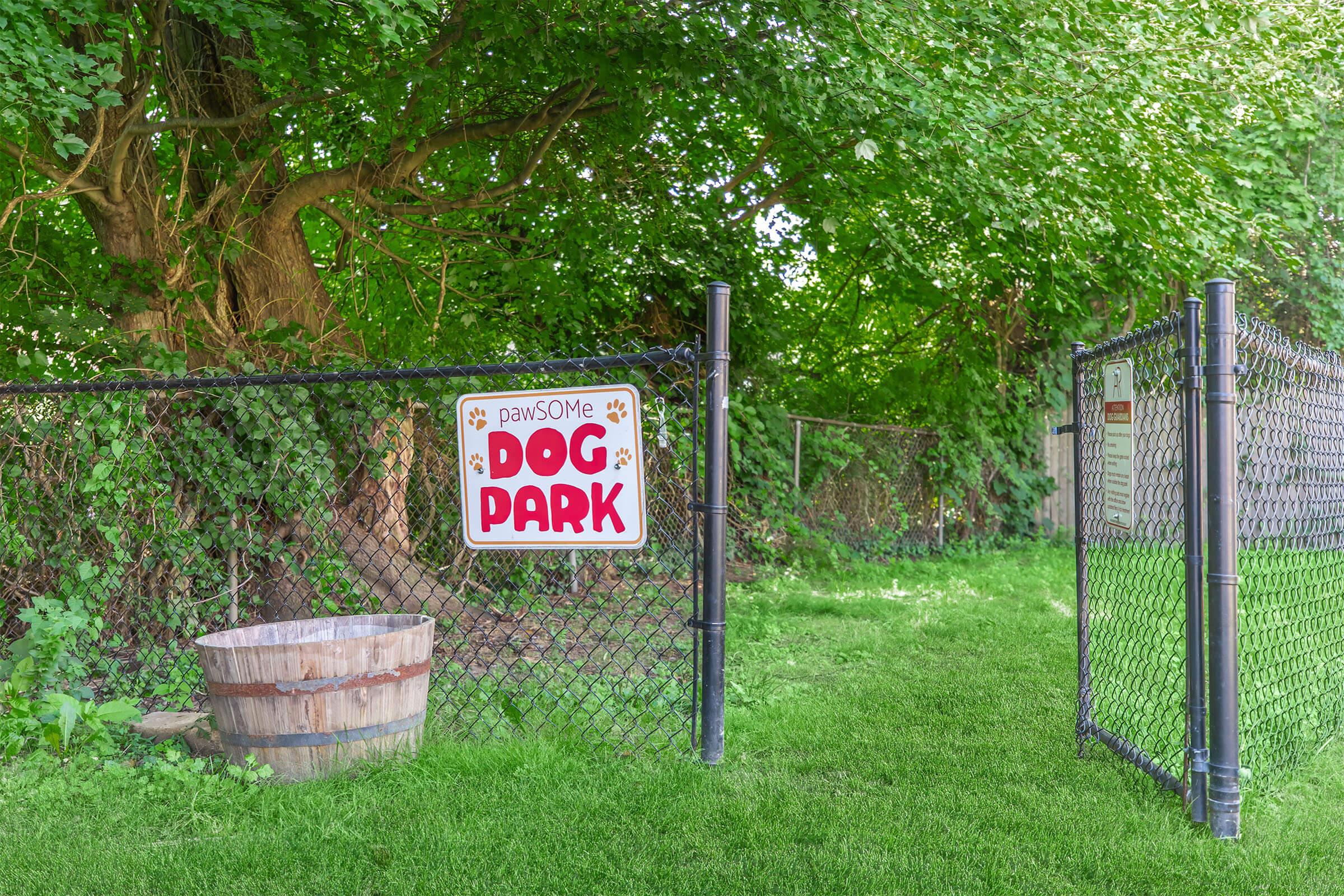 A sign reading "pawsome DOG PARK" hangs on a black chain-link fence, surrounded by lush green foliage. A wooden barrel sits nearby, with a grassy area leading into the park. The entrance invites dogs and their owners to enjoy the space.