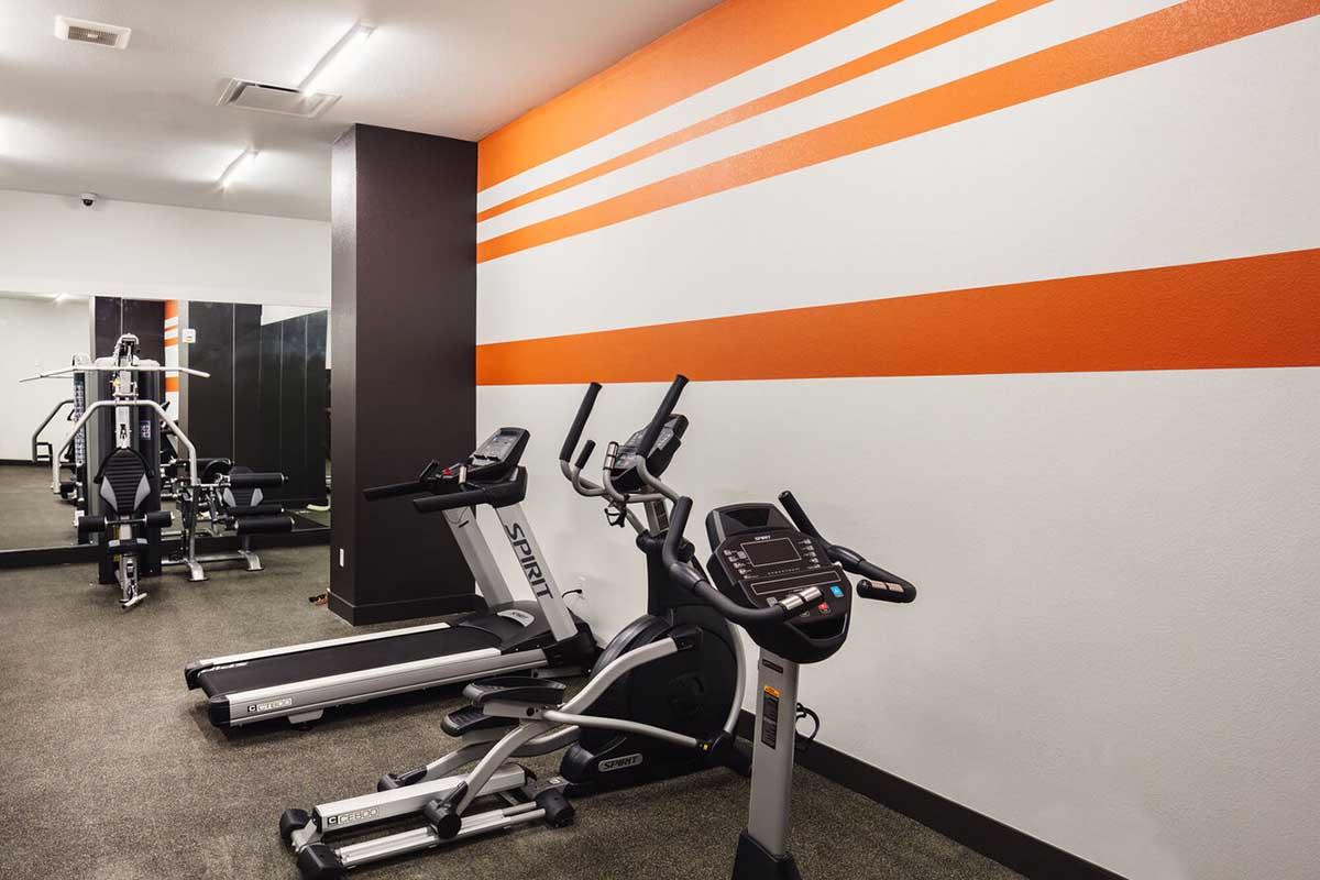 A modern gym space featuring exercise equipment, including a treadmill and an elliptical machine, against a backdrop of a textured wall with orange and white stripes. The floor is carpeted, and the environment is bright with overhead lighting.