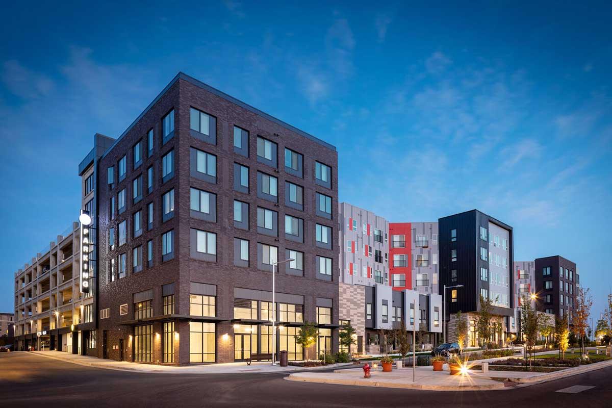 Modern multi-story buildings with a mix of brick and colorful facades. The architecture features large windows and a well-lit entrance area. Surrounding the buildings is a landscaped area with trees and paved walkways, set against a clear blue sky.