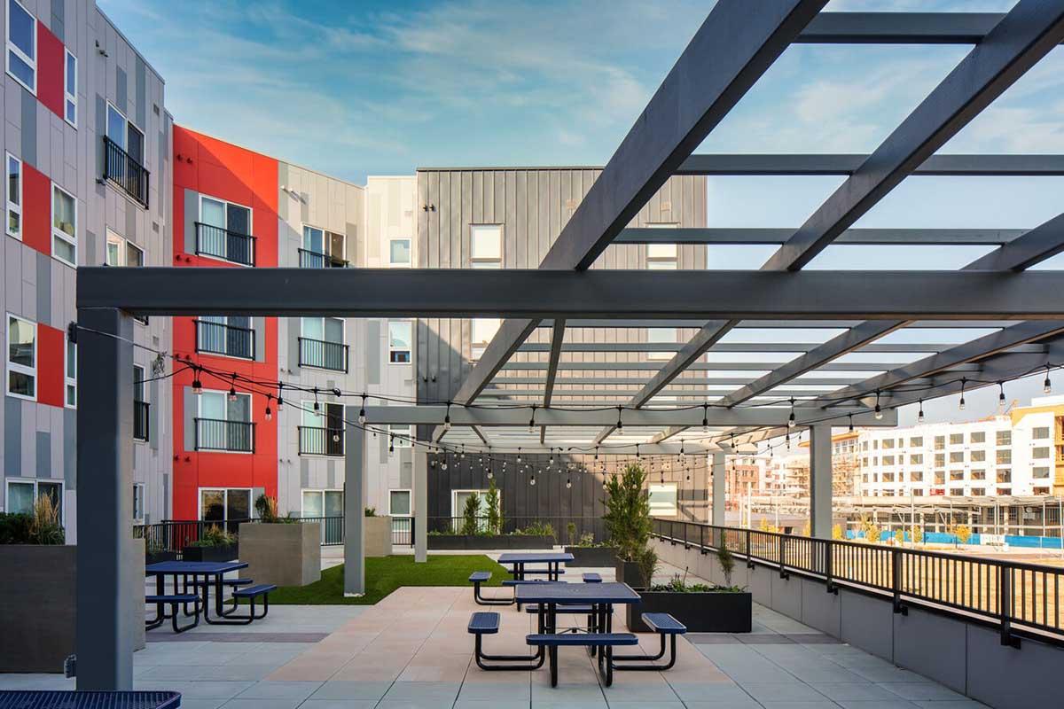 A modern outdoor space featuring a covered pergola with string lights, picnic tables, and green grass. The background showcases colorful apartment buildings and a blue sky, creating a vibrant and inviting atmosphere for social gatherings or relaxation.