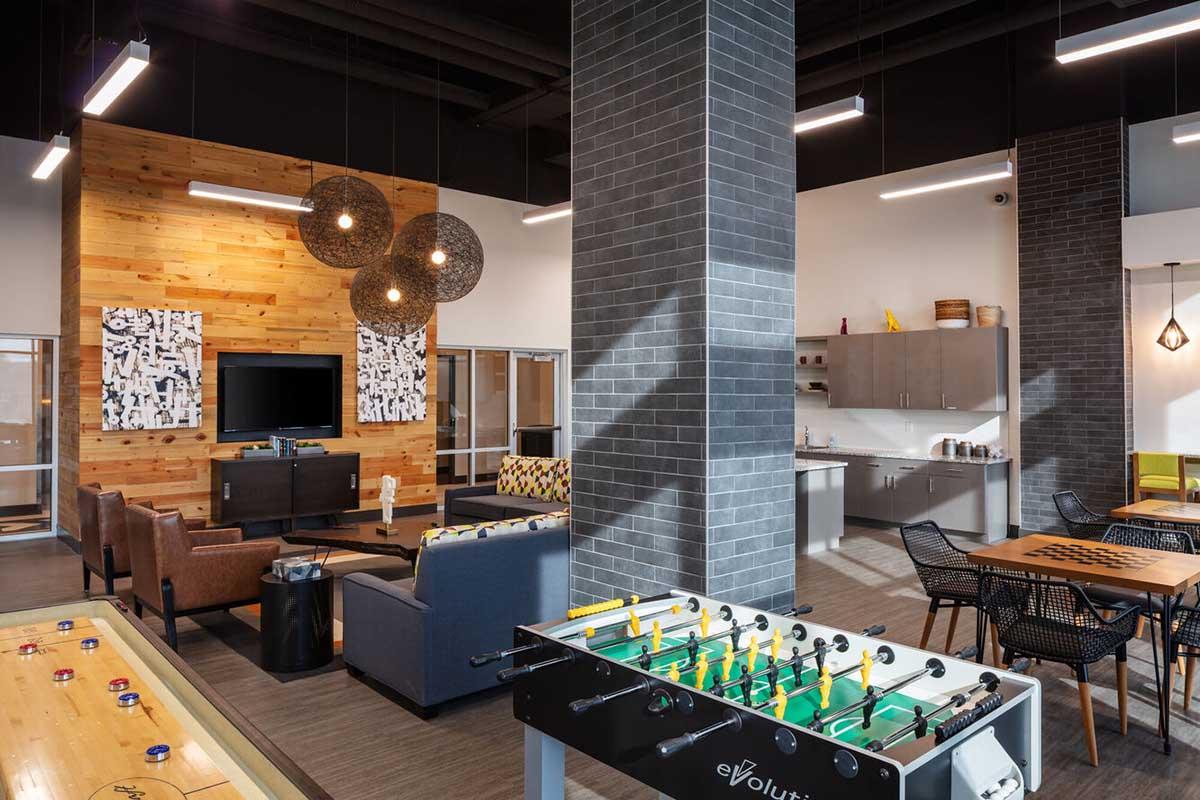 A modern lounge area featuring a foosball table, comfortable seating, a large TV, and a kitchenette. The decor includes wooden and grey elements, pendant lighting, and wall art, creating a welcoming atmosphere for socializing and relaxation.