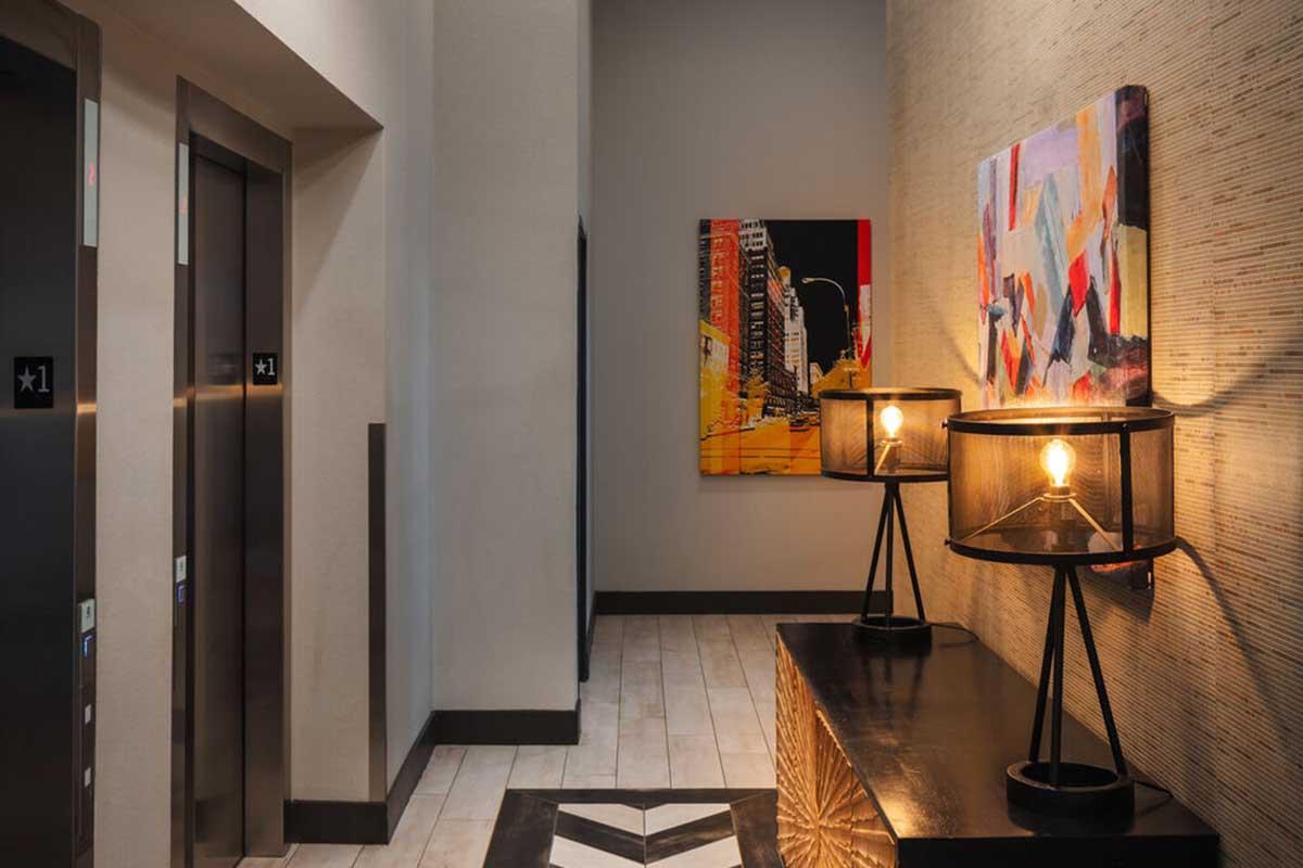 A modern hallway featuring two elevator doors and a decorative wall. On one side, there are two stylish lamps on a black table, providing warm lighting. A colorful abstract painting hangs on the wall, adding an artistic touch to the contemporary setting. The floor is tiled with light wood planks.