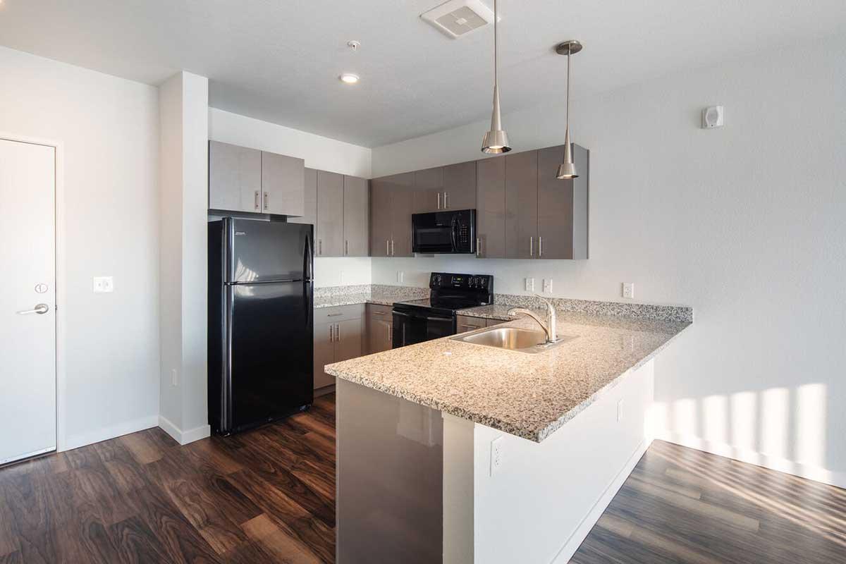 Modern kitchen featuring granite countertops, stainless steel appliances including an oven, microwave, and refrigerator, sleek cabinets, and wooden flooring. Large windows allow natural light to brighten the space.