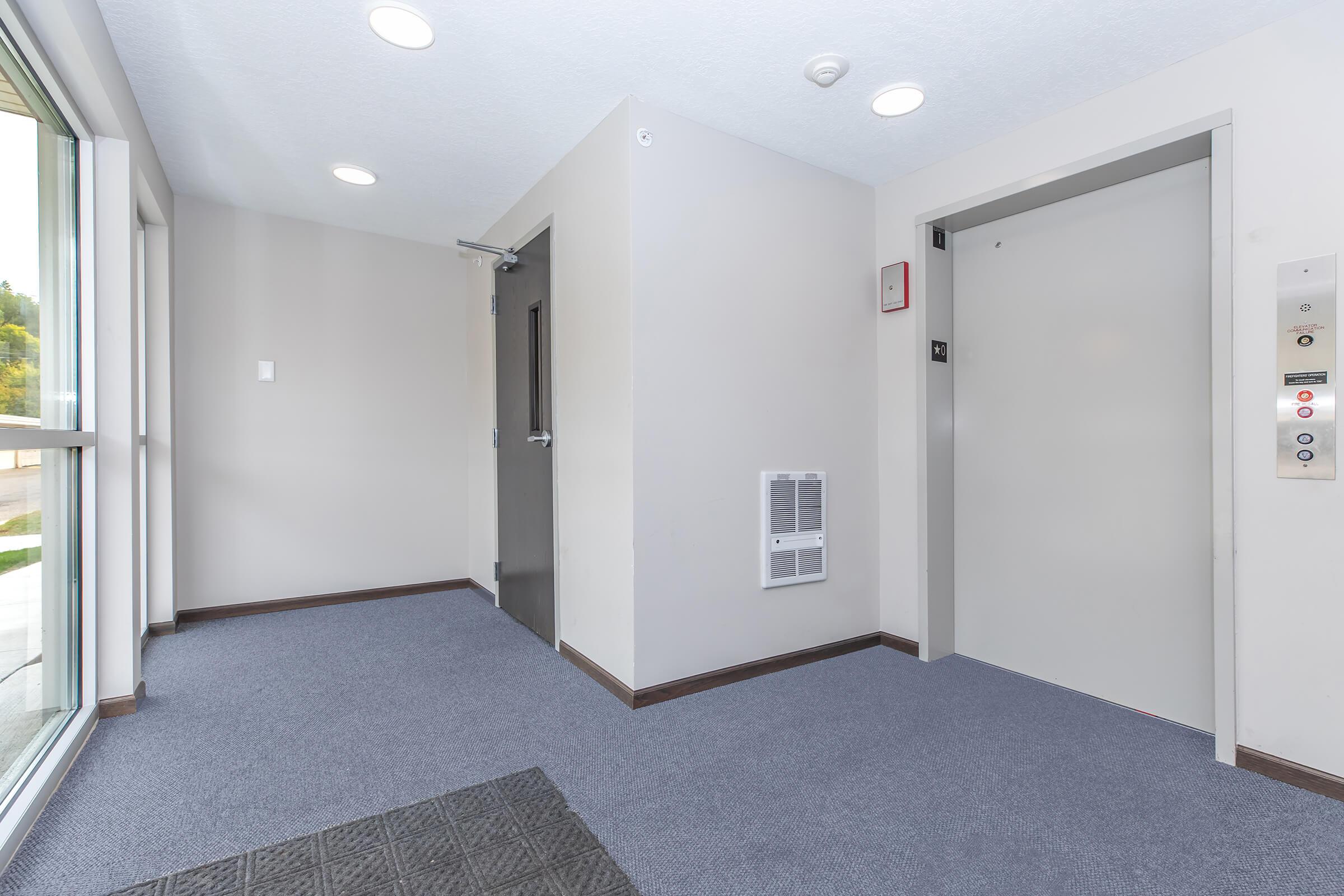 A well-lit interior hallway with gray walls and carpeting. There is a closed elevator door on the right, with elevator controls next to it. To the left, there is a gray wall with a door and a security feature. Large windows provide natural light, creating a modern and inviting atmosphere.