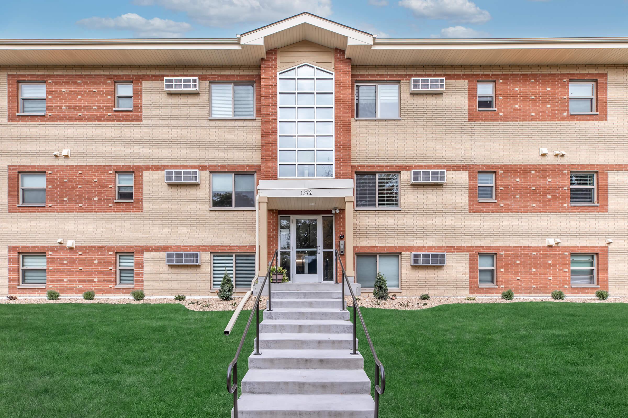 A three-story apartment building featuring a combination of red brick and beige siding. The main entrance has a large glass canopy, with stairs leading up to it. Air conditioning units are visible on the windows. A neatly maintained lawn surrounds the building.
