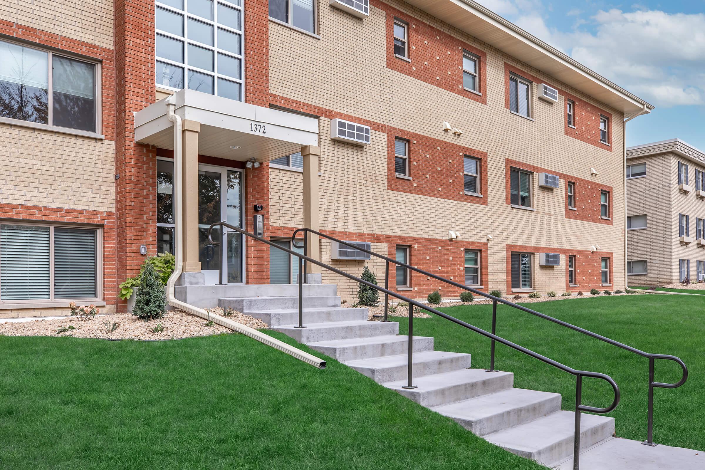 Exterior view of a multi-story apartment building featuring a brick facade, with prominent steps leading up to the entrance. The entrance includes a covered awning and glass doors, and the landscaped area features well-maintained grass and small shrubs. Air conditioning units are visible on the windows.