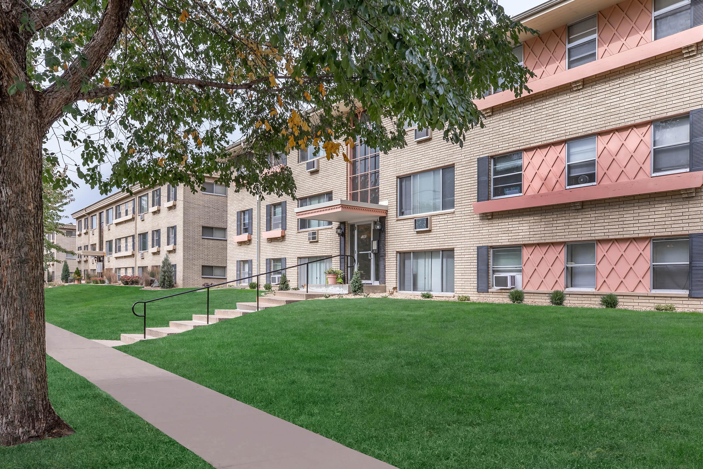 A view of a low-rise brick apartment building with a landscaped front yard and a tree nearby. The building features a mix of window styles and colored accents. A wide pathway leads to the entrance, and there are small shrubs along the building's facade. The sky is overcast, indicating a cloudy day.