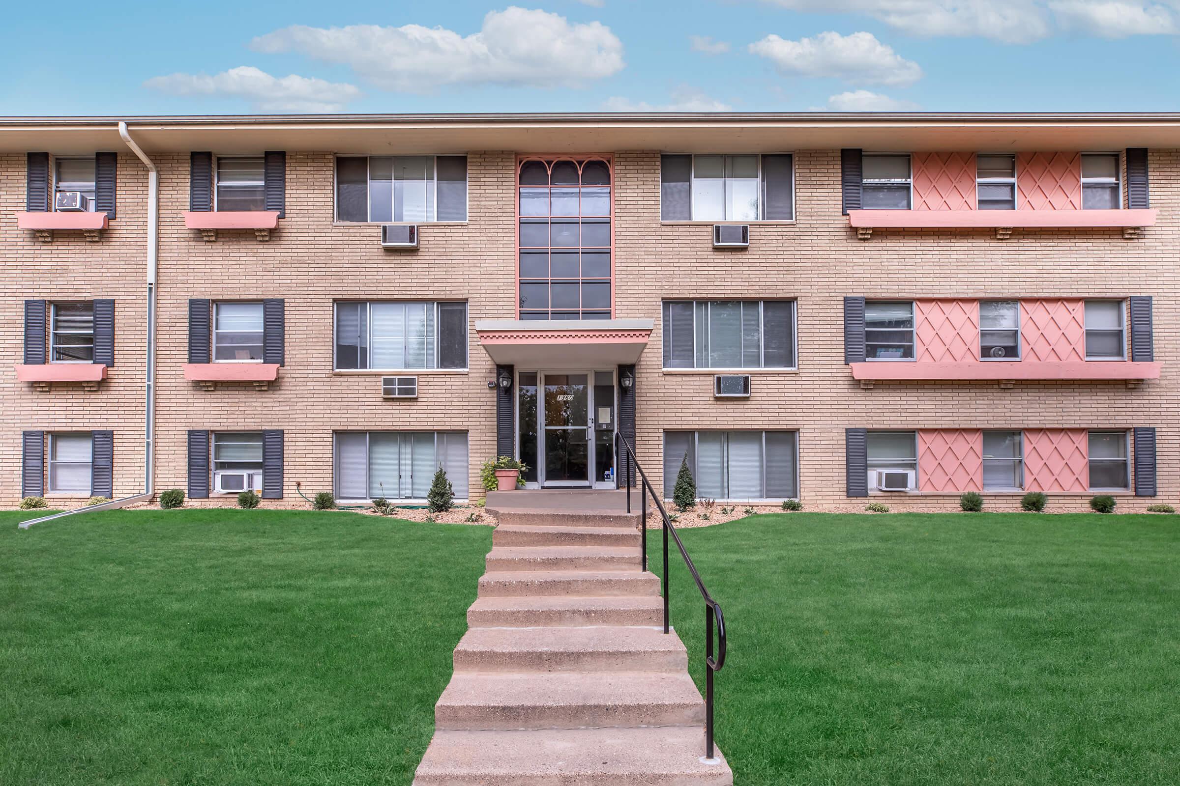 A three-story apartment building with a brick facade, featuring large windows, a central entrance, and decorative pink accents on the balconies. A set of steps leads up to the entrance, surrounded by well-maintained grass and small plants. The sky is clear with a few clouds visible.