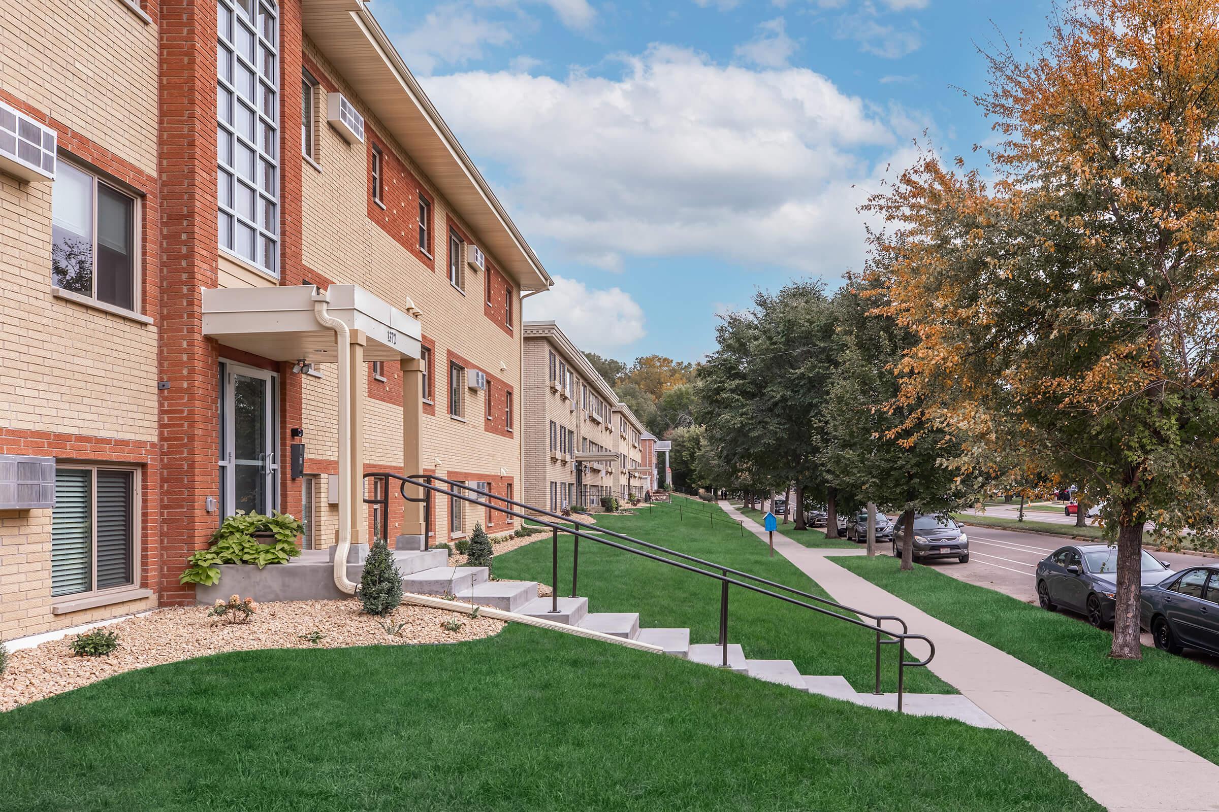A well-maintained residential building with brick exterior and large windows. There are landscaped lawns with shrubs and small trees, and a pathway leading to the entrance. The sidewalk runs along the street lined with parked cars, under a partly cloudy sky.