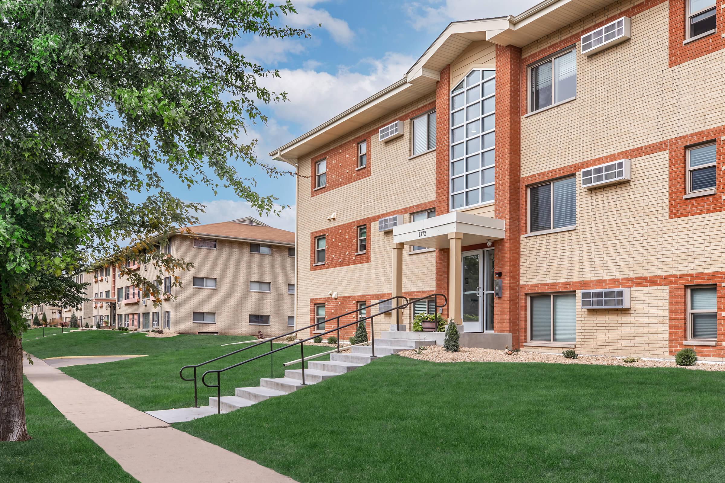 A modern multi-unit apartment building with a brick facade, featuring a stairway leading to the entrance. The building has large windows and air conditioning units. Lush green grass and a pathway are visible in the foreground, with additional apartment buildings in the background under a partly cloudy sky.