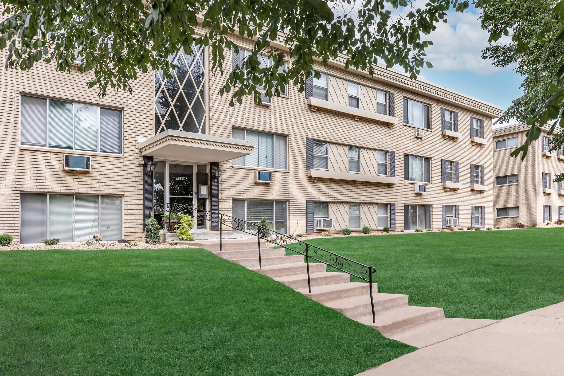 A multi-story brick apartment building featuring large windows, air conditioning units, and a decorative front entrance. The building is set against a well-manicured lawn with green grass and a few small shrubs. A set of concrete stairs leads up to the entrance.