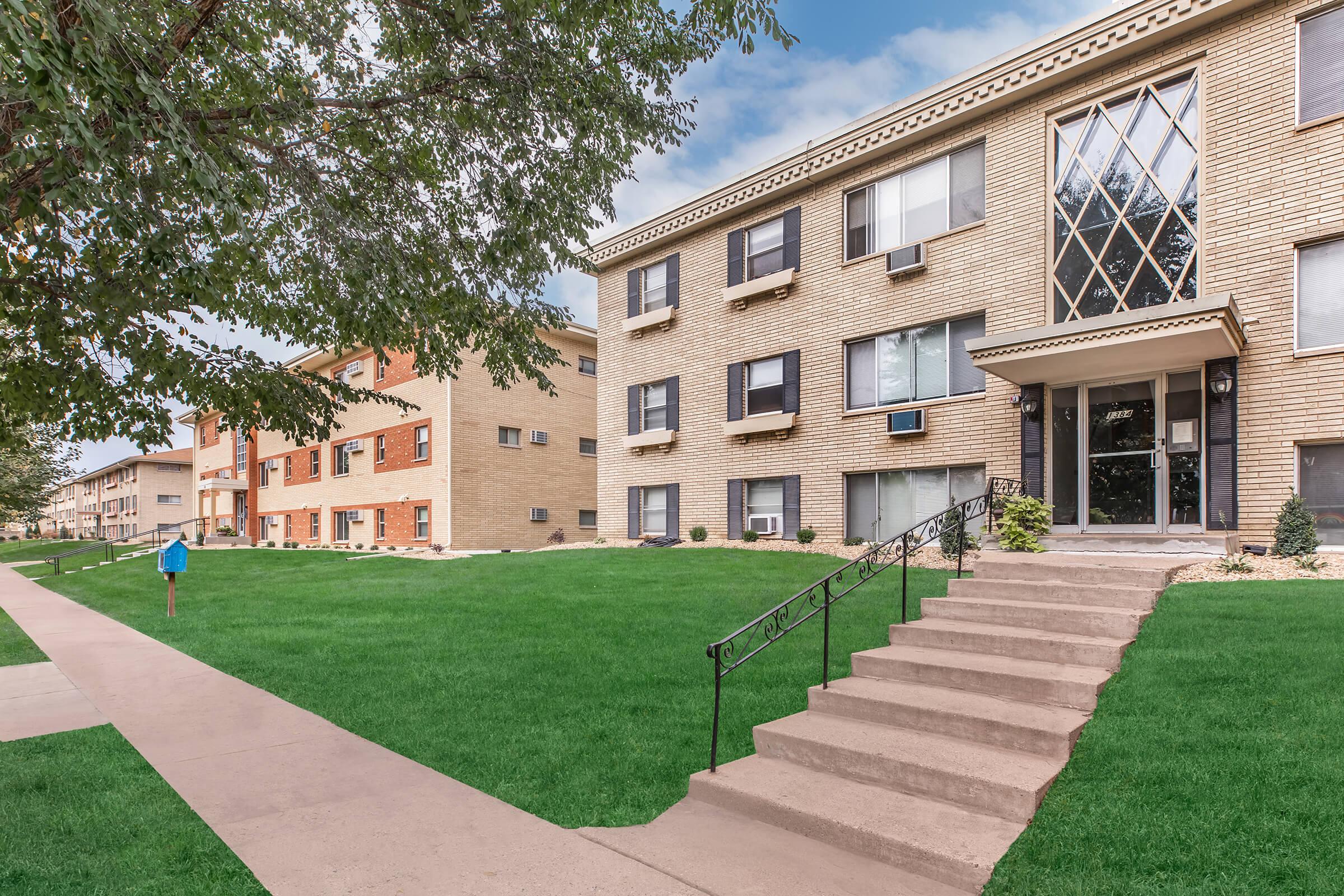 A residential building with a brick exterior, featuring several windows and air conditioning units. The entrance includes a set of stairs leading to a glass door. Lush green lawns surround the building, and a sidewalk runs parallel to the front. In the background, there are additional buildings with similar architectural styles.