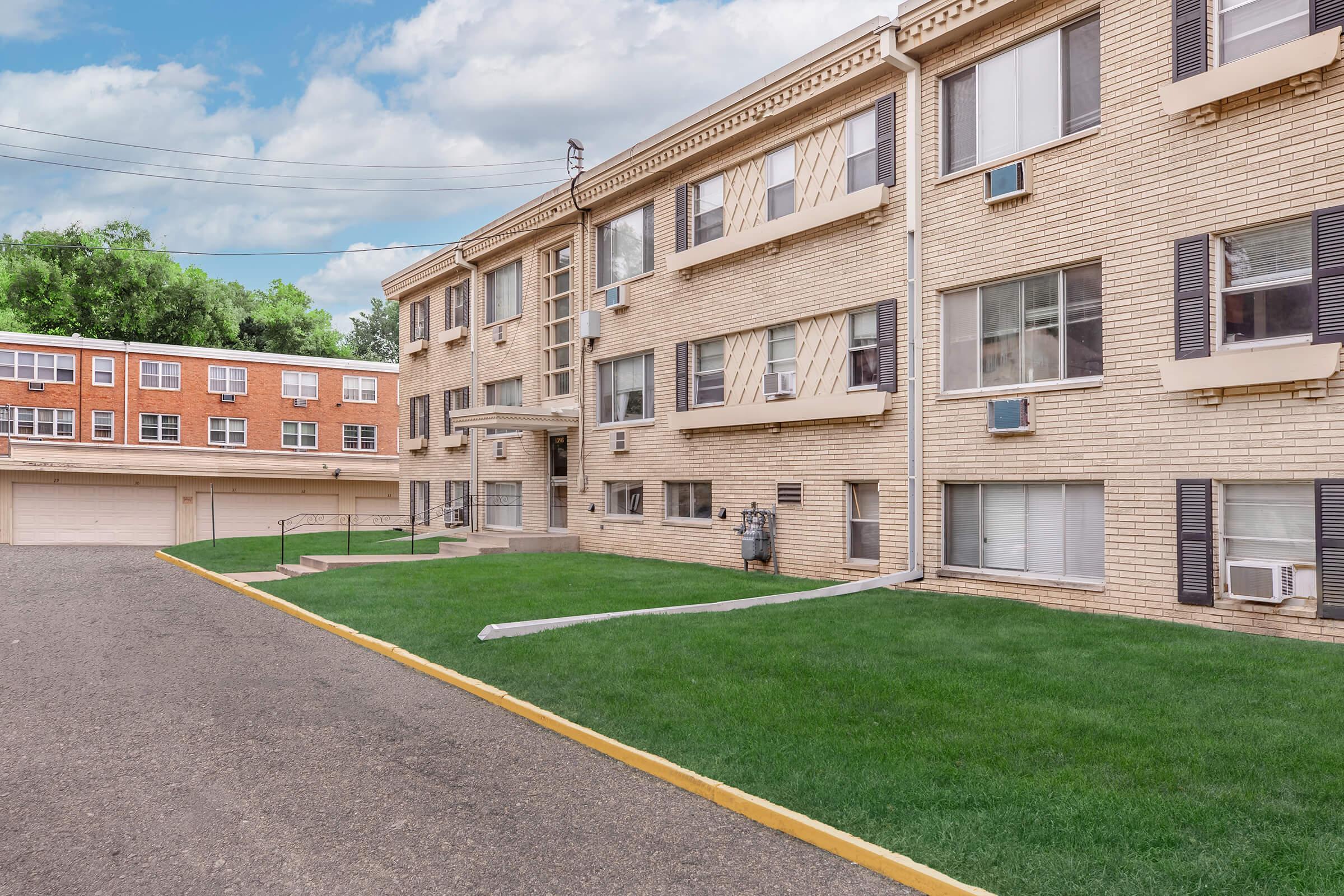 A view of a brick apartment building with multiple windows and air conditioning units, surrounded by well-manicured green lawns. The building features a small set of stairs leading to the entrance, with a driveway and another building visible in the background under a partly cloudy sky.