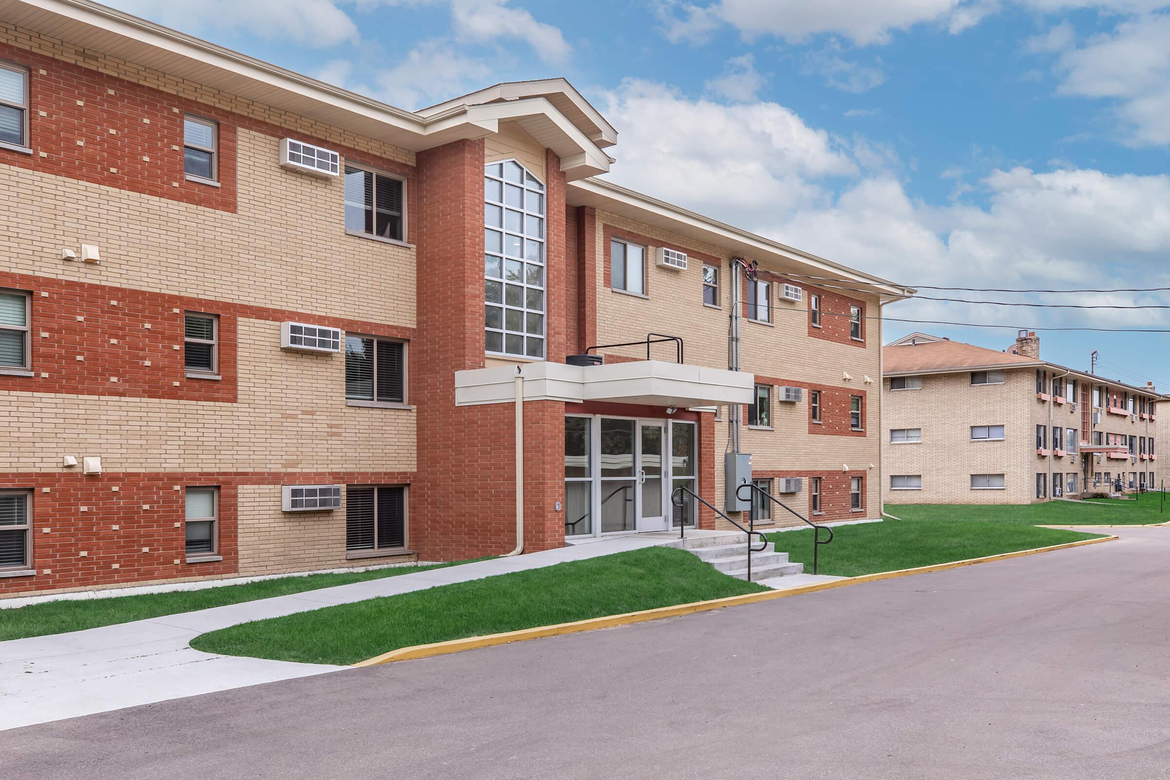 A modern brick apartment building featuring a glass entrance and accessible stairs, with green grass surrounding the property. The exterior includes various windows, and nearby, there are additional buildings visible along the street. The sky is partly cloudy, suggesting a pleasant day.