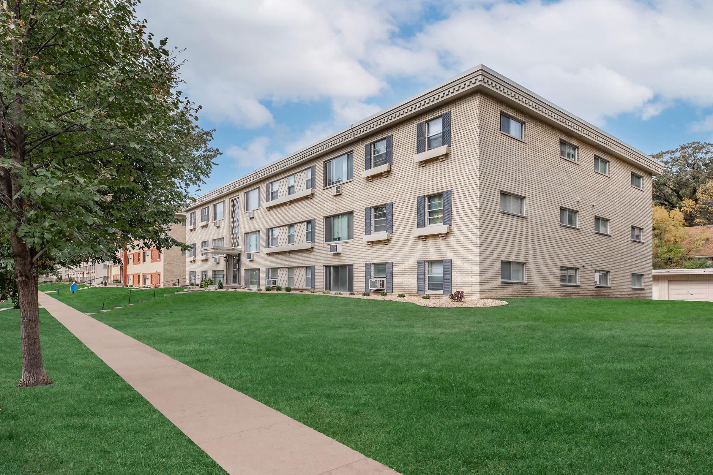 A three-story brick apartment building with multiple windows and air conditioning units, surrounded by a well-maintained lawn. A pathway runs along the front of the building, leading to a landscaped area. Trees are visible nearby, and the sky is partly cloudy.