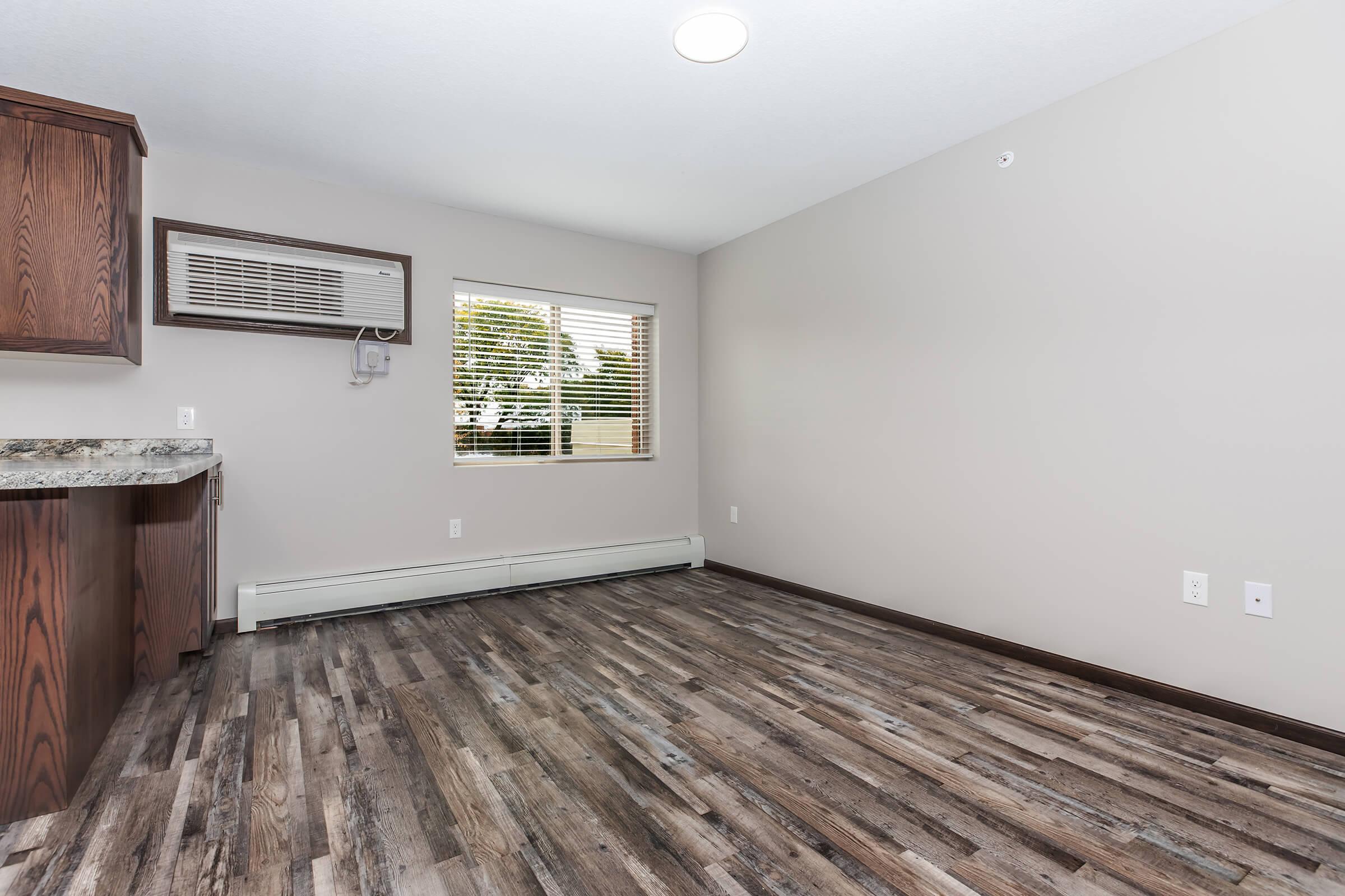 A spacious, empty room featuring light brown wood-like flooring, beige walls, and a large window with blinds. There’s an air conditioning unit on the wall and a kitchen area with dark wooden cabinets. The overall atmosphere is bright and uncluttered, suitable for various furniture arrangements.