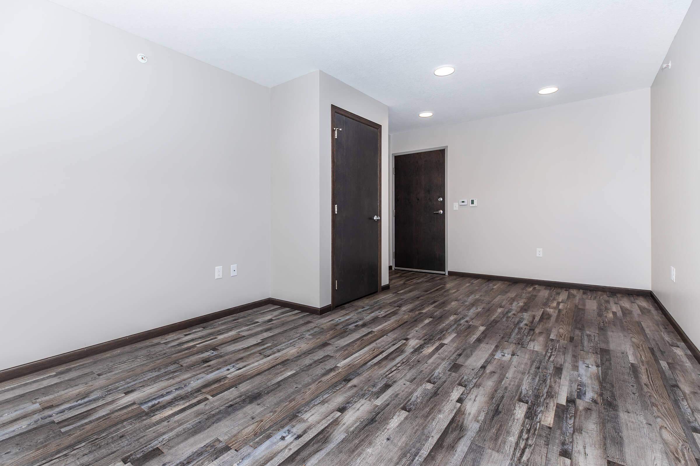 A modern, empty room featuring light-colored walls, dark wood doors, and a laminate floor with a wood finish. There are two doors on opposite walls and recessed lighting in the ceiling, creating a bright and spacious appearance. The room is designed for versatility and potential furnishing.