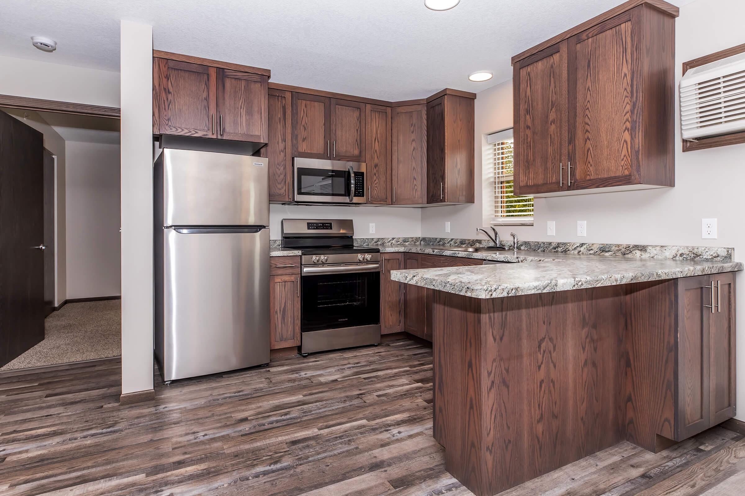 Modern kitchen featuring dark wood cabinetry, stainless steel appliances, a countertop with a marbled design, and a spacious layout. The kitchen includes a refrigerator, oven, microwave, and sink, with a view of an adjacent room and natural light coming through the window. The flooring is a wood-like laminate.