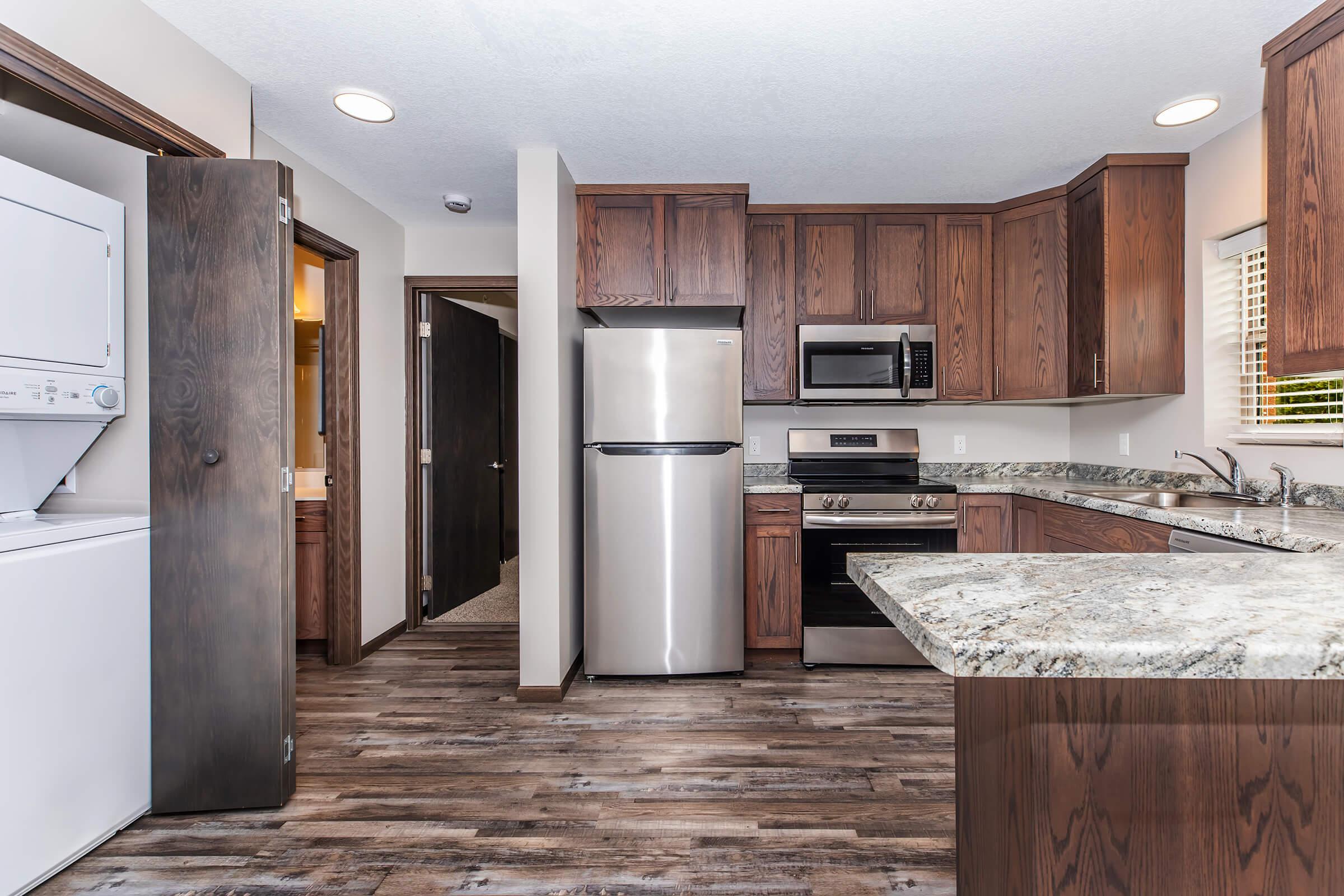 A modern kitchen with dark wooden cabinets, a stainless steel refrigerator, a stove, and granite countertops. The space features a washer and dryer in a separate area and has a warm, inviting atmosphere with natural light coming through a window. The flooring is a dark laminate that complements the cabinetry.