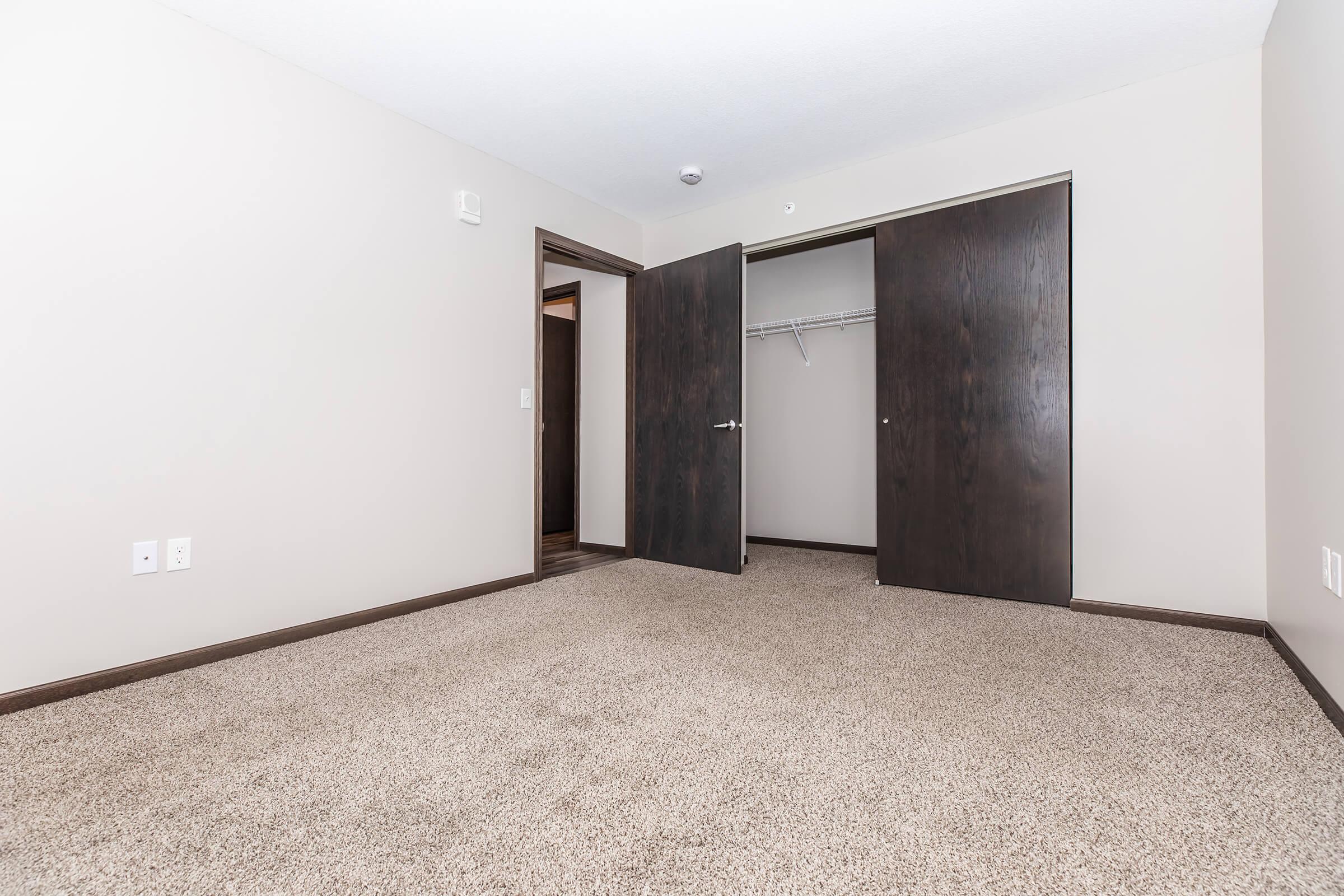 A spacious, empty room featuring light-colored walls and a plush carpet. Two dark wooden closet doors are open, revealing an empty closet space. The room has a single door leading to another area, providing a simple and modern appearance ideal for various purposes.
