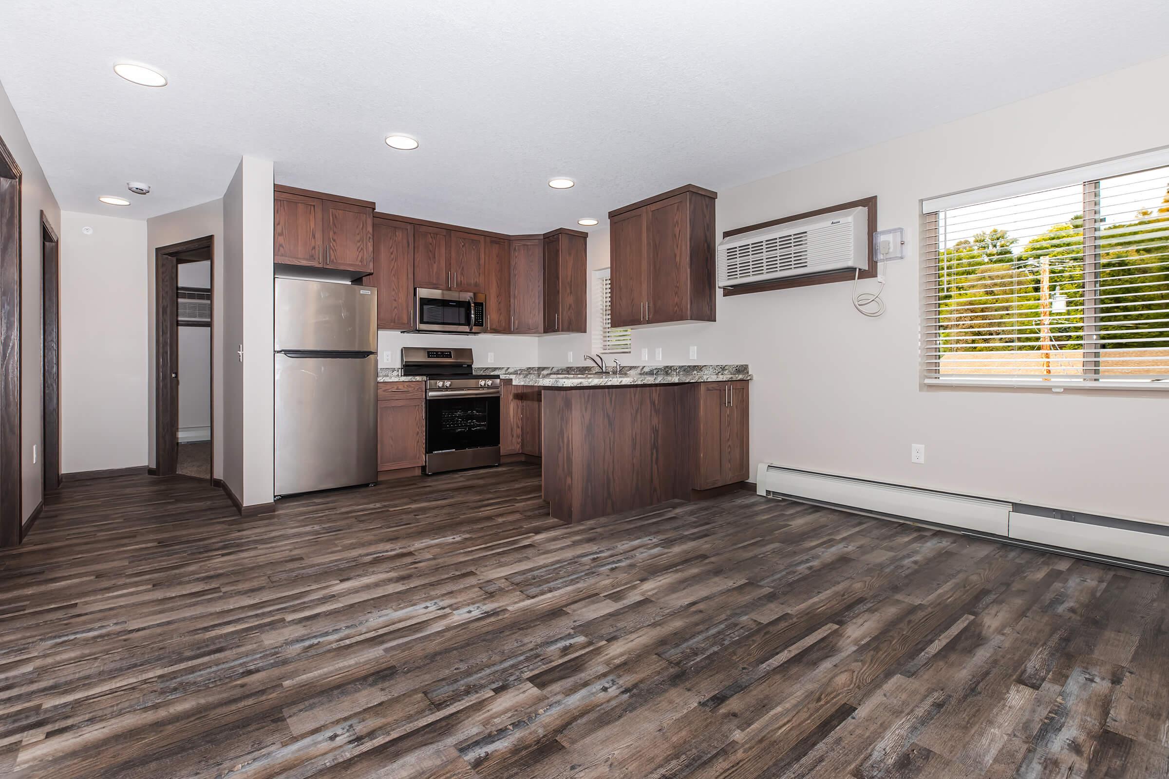 Modern kitchen and living area featuring dark wood cabinets, stainless steel appliances, and a countertop. Includes an open layout with hardwood flooring and large windows allowing natural light. Air conditioning unit mounted on the wall, with an inviting atmosphere for dining and living.