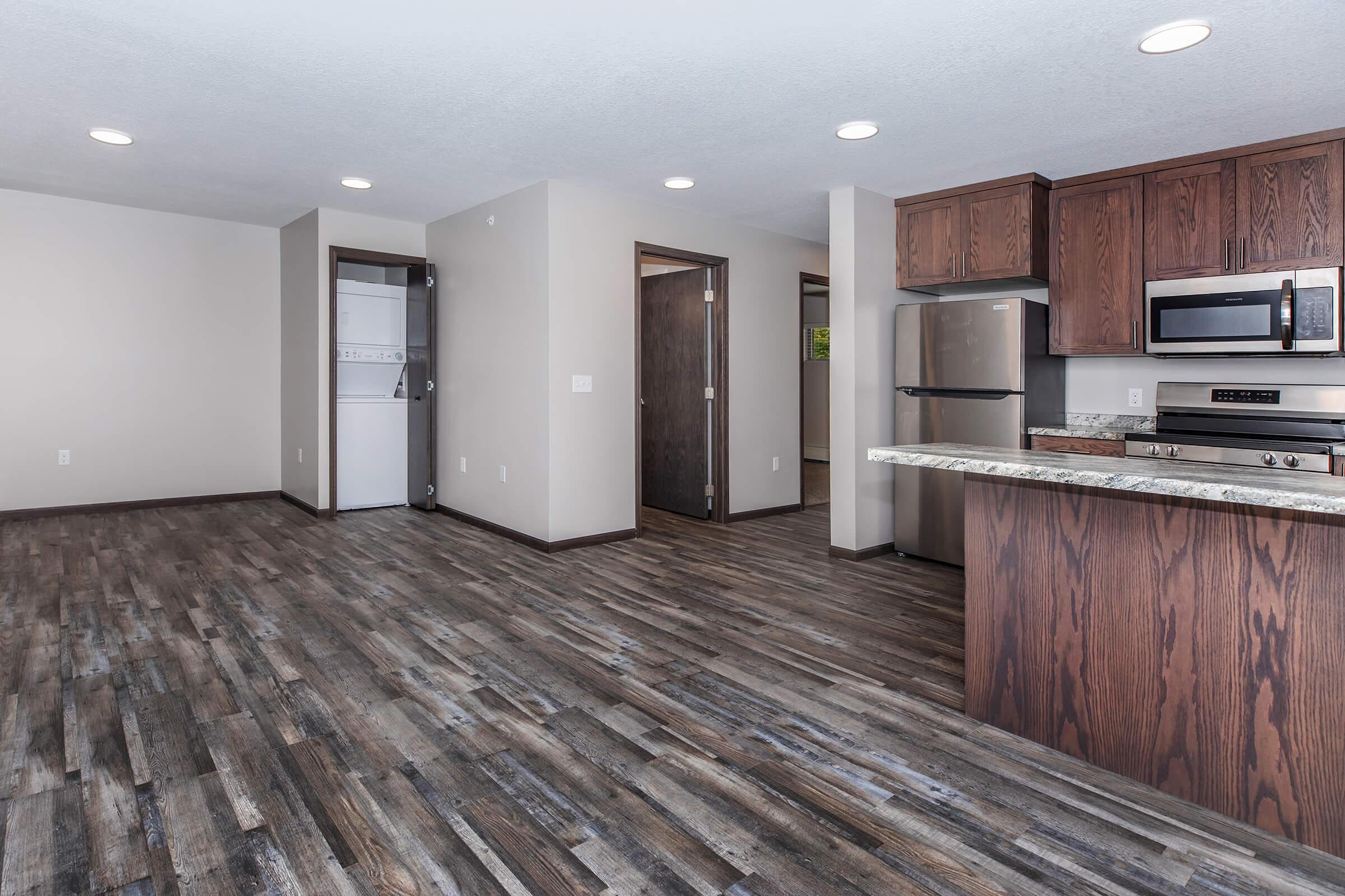 Modern, spacious living area with dark wood cabinetry and stainless steel appliances. The kitchen includes a countertop and connects to an open living space with laminate flooring. A laundry area is visible in the background, along with two doorways leading to separate rooms. Well-lit with recessed lighting.