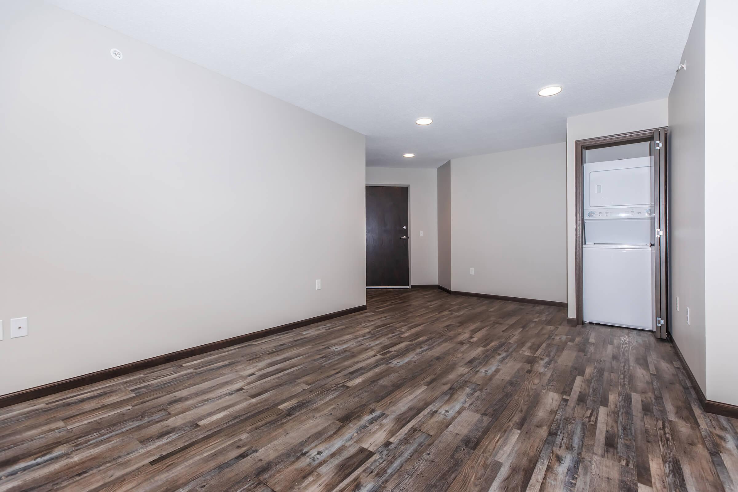 A spacious, unfurnished apartment interior featuring light-colored walls and dark wood flooring. There are visible appliances in a closet on the right, and a doorway leading to another room or entrance in the background. Natural light illuminates the area from recessed ceiling lights.
