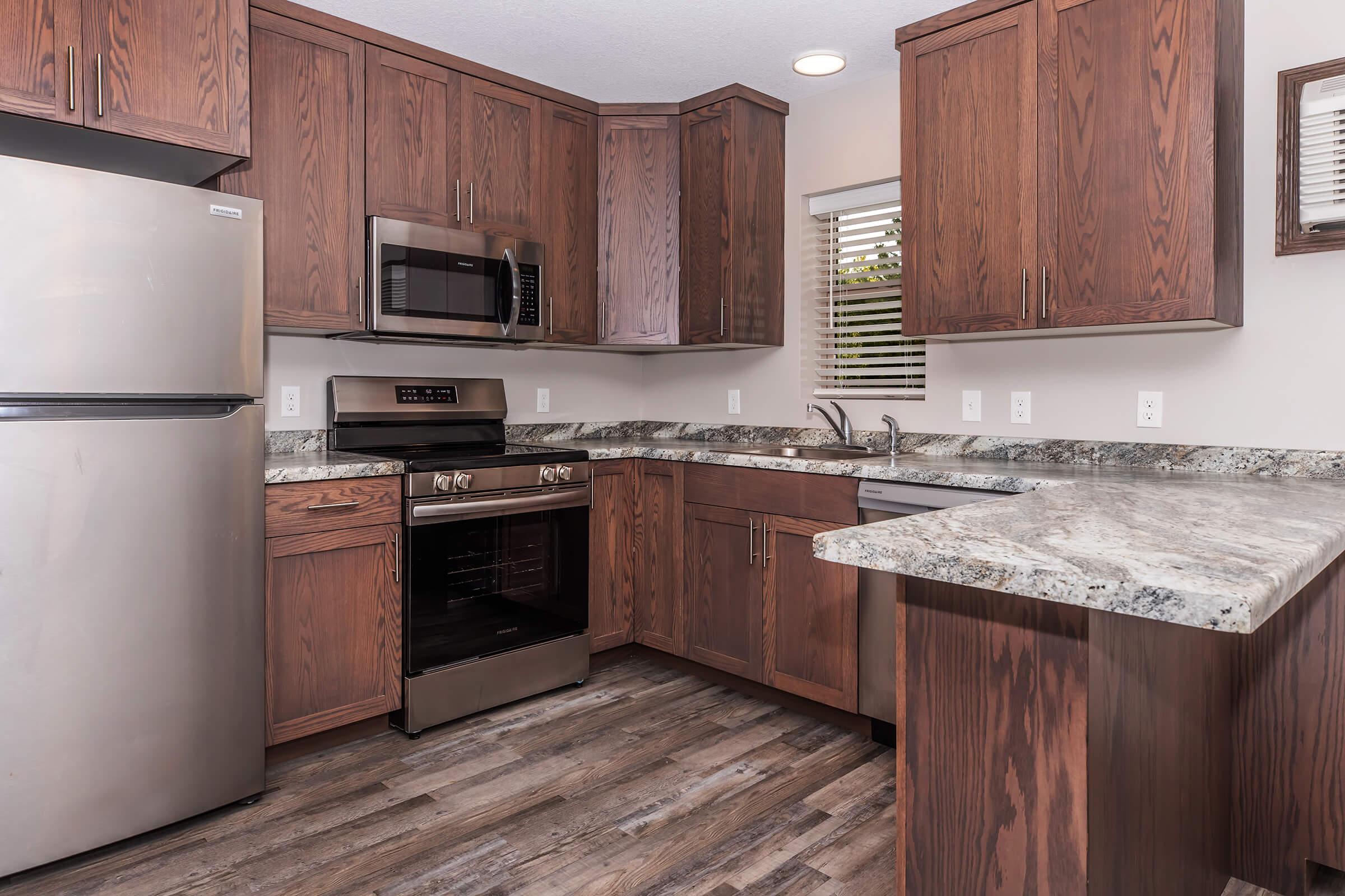 Modern kitchen featuring dark wood cabinets, stainless steel appliances including a fridge, microwave, and oven. The countertop is a light granite finish, with a sink under a window. The floor is a wood-like laminate, creating a warm and inviting atmosphere.