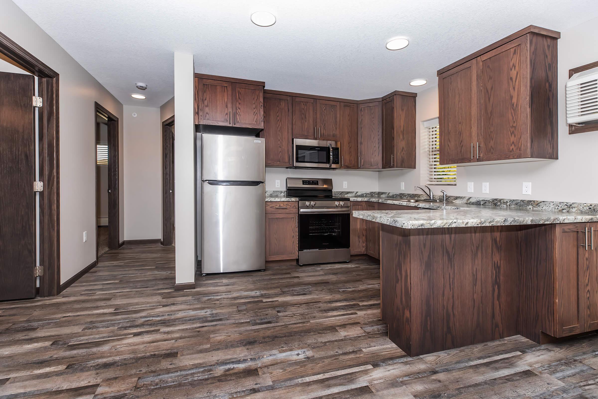 Modern kitchen featuring dark wood cabinetry, stainless steel appliances including a fridge and microwave, and a stylish granite countertop. The open layout includes a dining area, light hardwood flooring, and recessed lighting, creating a warm and inviting atmosphere.