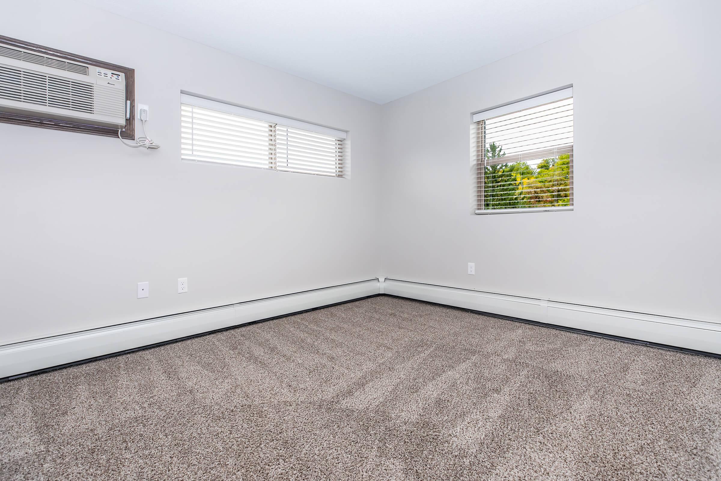 A spacious, empty room featuring light gray walls and large windows with blinds. An air conditioning unit is mounted on the wall, and the floor is covered with plush, beige carpeting. Natural light fills the space, highlighting its clean and simple design, ideal for various uses or decoration.
